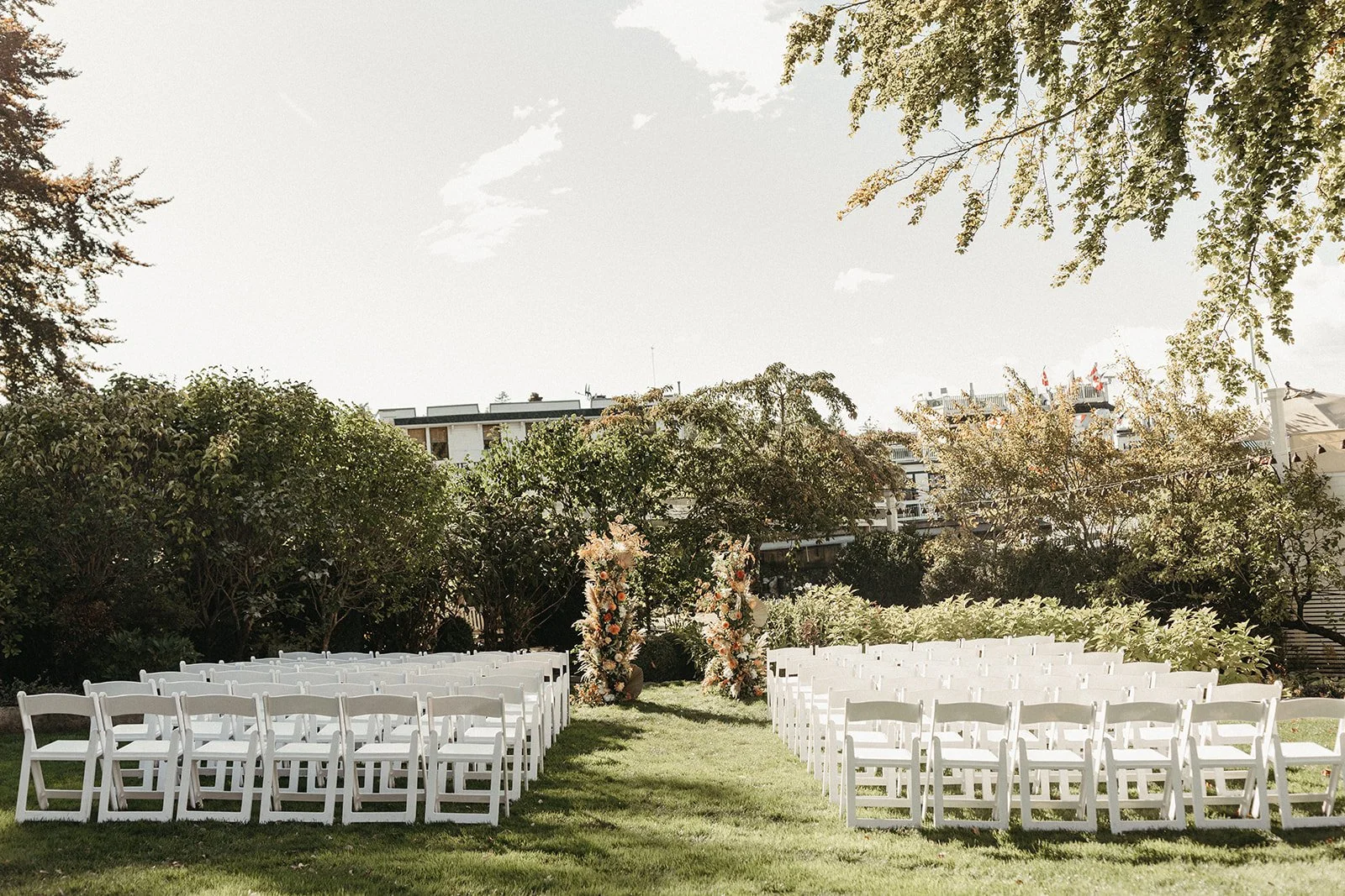 Outdoor wedding ceremony setup with white chairs arranged on grass, floral arch, surrounded by trees and bushes, in a park or garden setting.