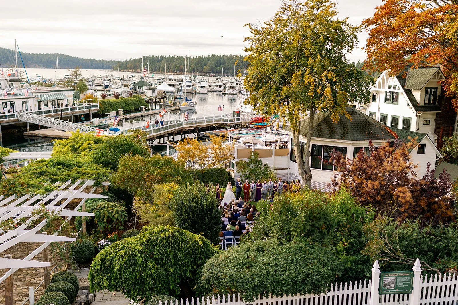 A wedding ceremony taking place outdoors near a marina with boats, trees with fall foliage, and a white house in the background.