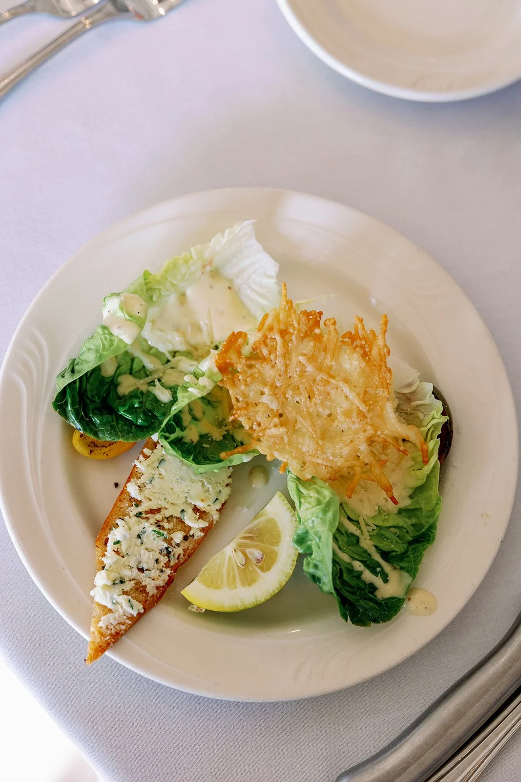 Appetizer plate with salad, lemon wedge, a slice of bread topped with cheese and herbs, and a fried onion ring.