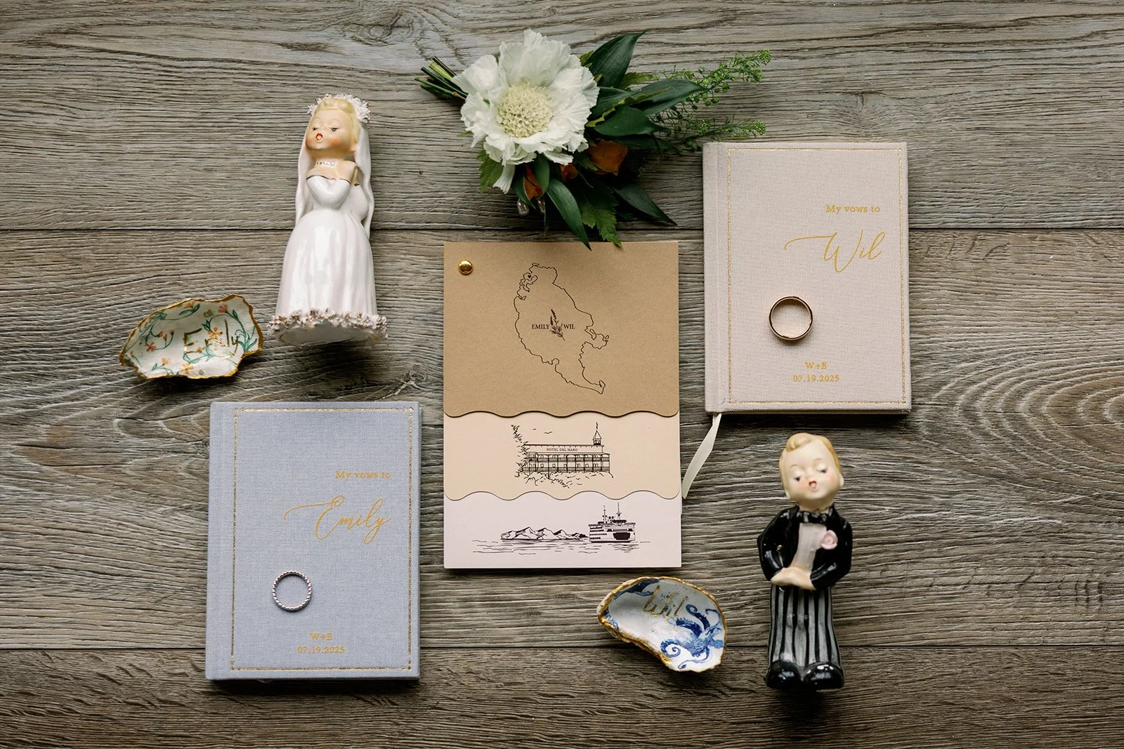 Wedding rings, figurines of a bride and groom, floral bouquet, decorative ceramic shells, and wedding vow books on a wooden surface.