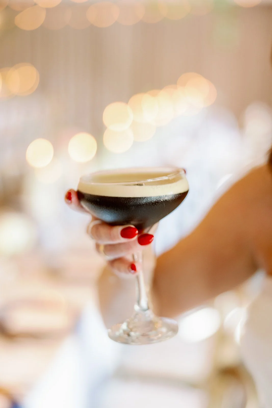 Person holding a dark cocktail with a frothy top in a coupe glass, with red painted nails, at a festive event with blurred lights in the background.
