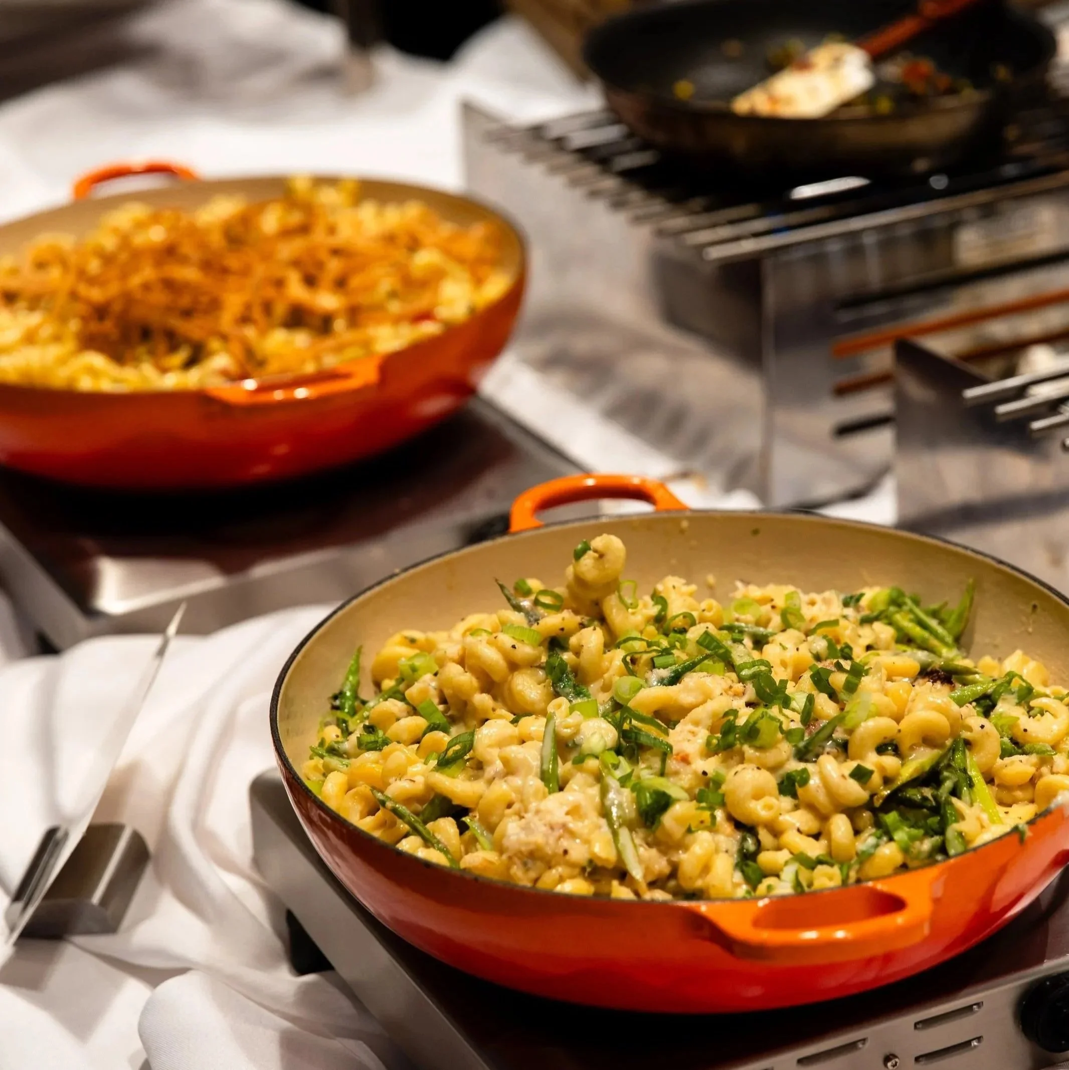 Two orange pots of cooked food, with one containing a mixture of corn, greens, and possibly chicken, and the other containing pasta with sauce, set on a buffet table.
