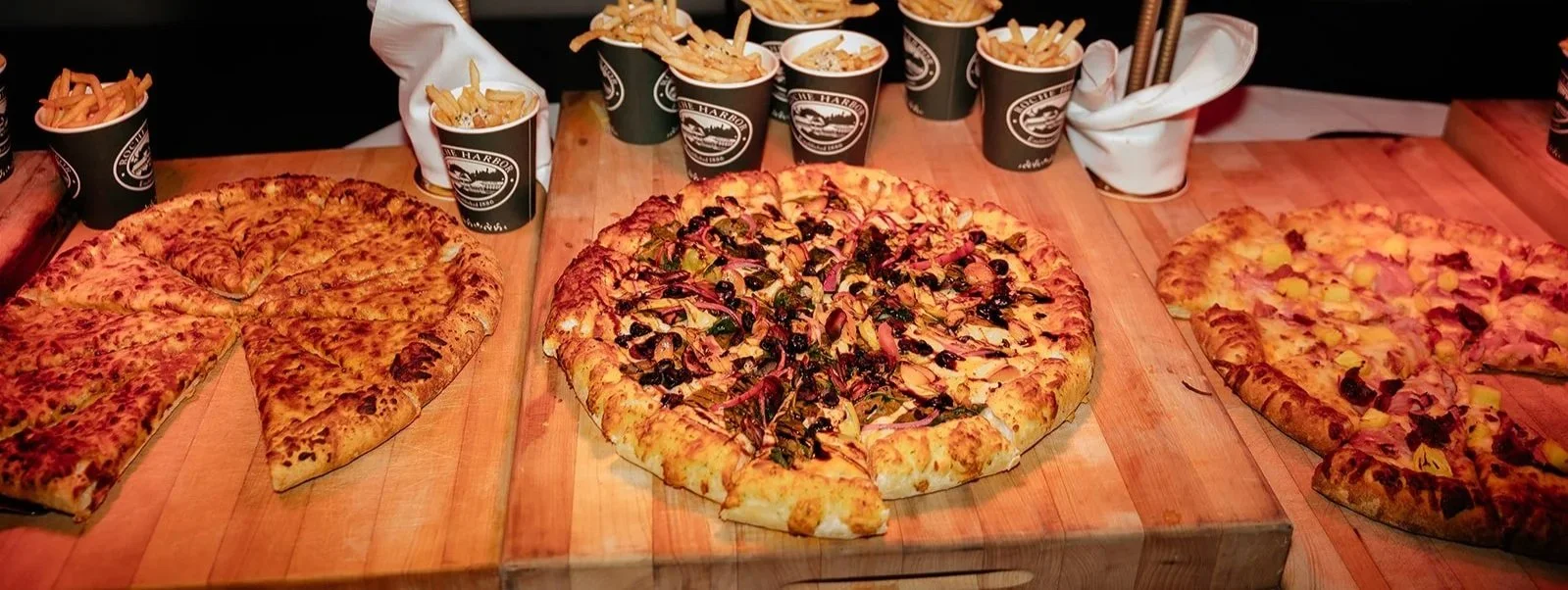 Three large pizzas and several cups of french fries on a wooden table.