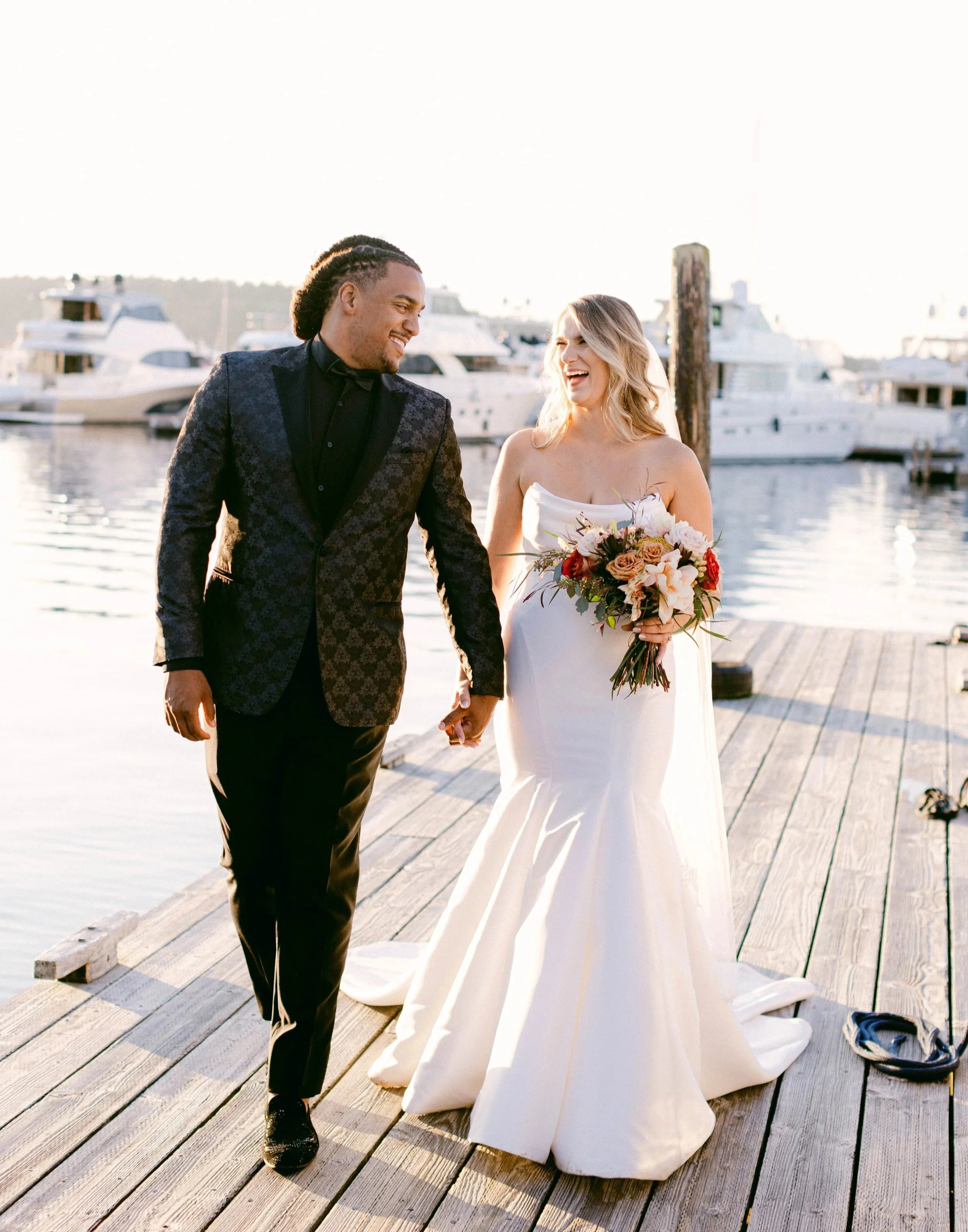 A bride and groom holding hands on a dock near boats, smiling at each other during their wedding at sunset.