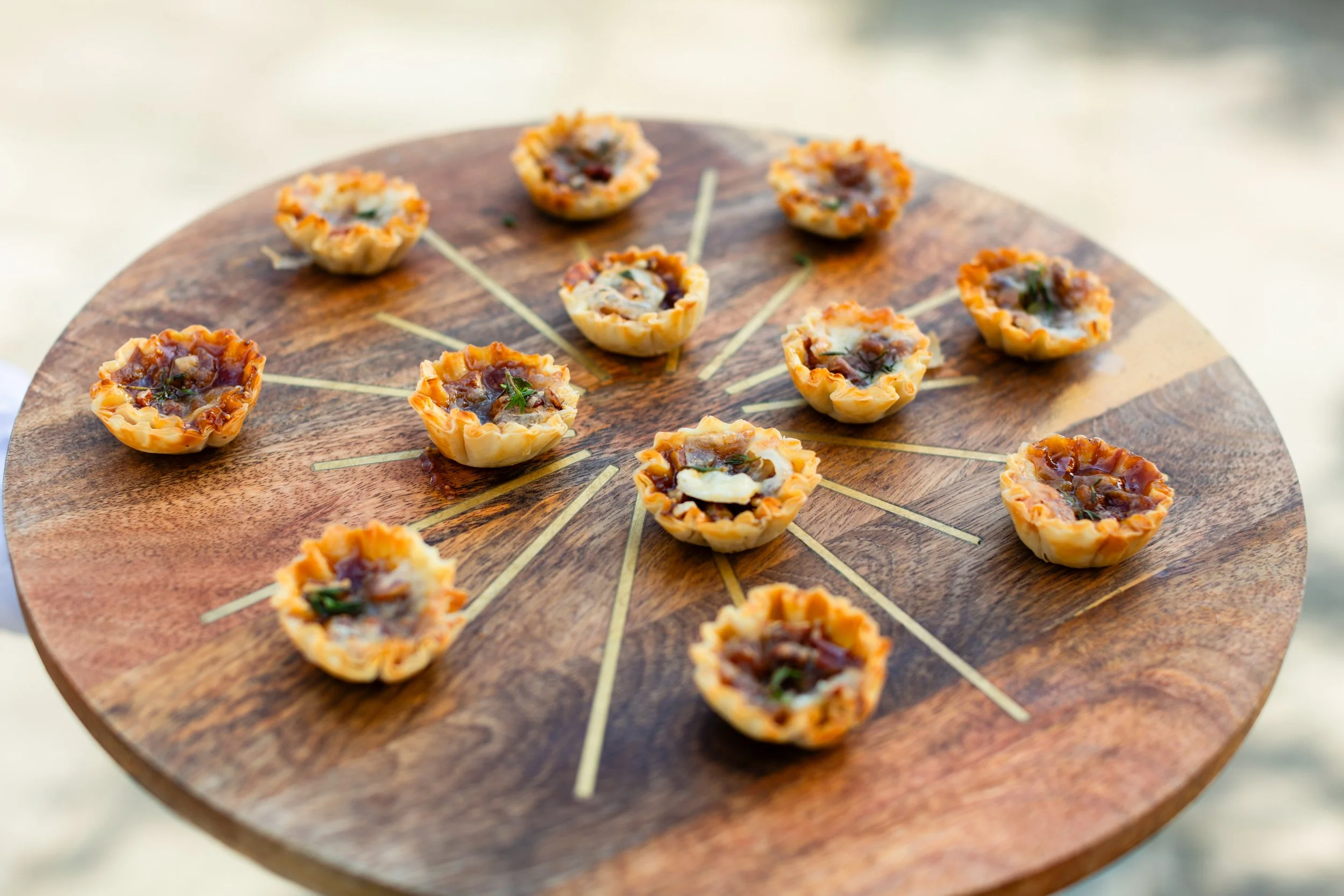 A round wooden serving board with small, cup-shaped savory appetizers topped with various ingredients.