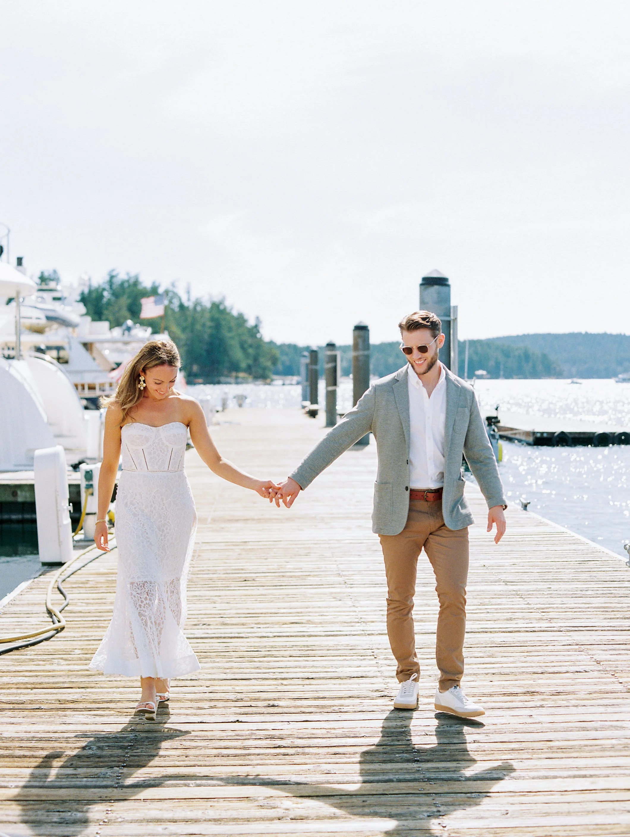 A couple walking hand in hand on a wooden dock by a marina with boats, water, and trees in the background on a sunny day.