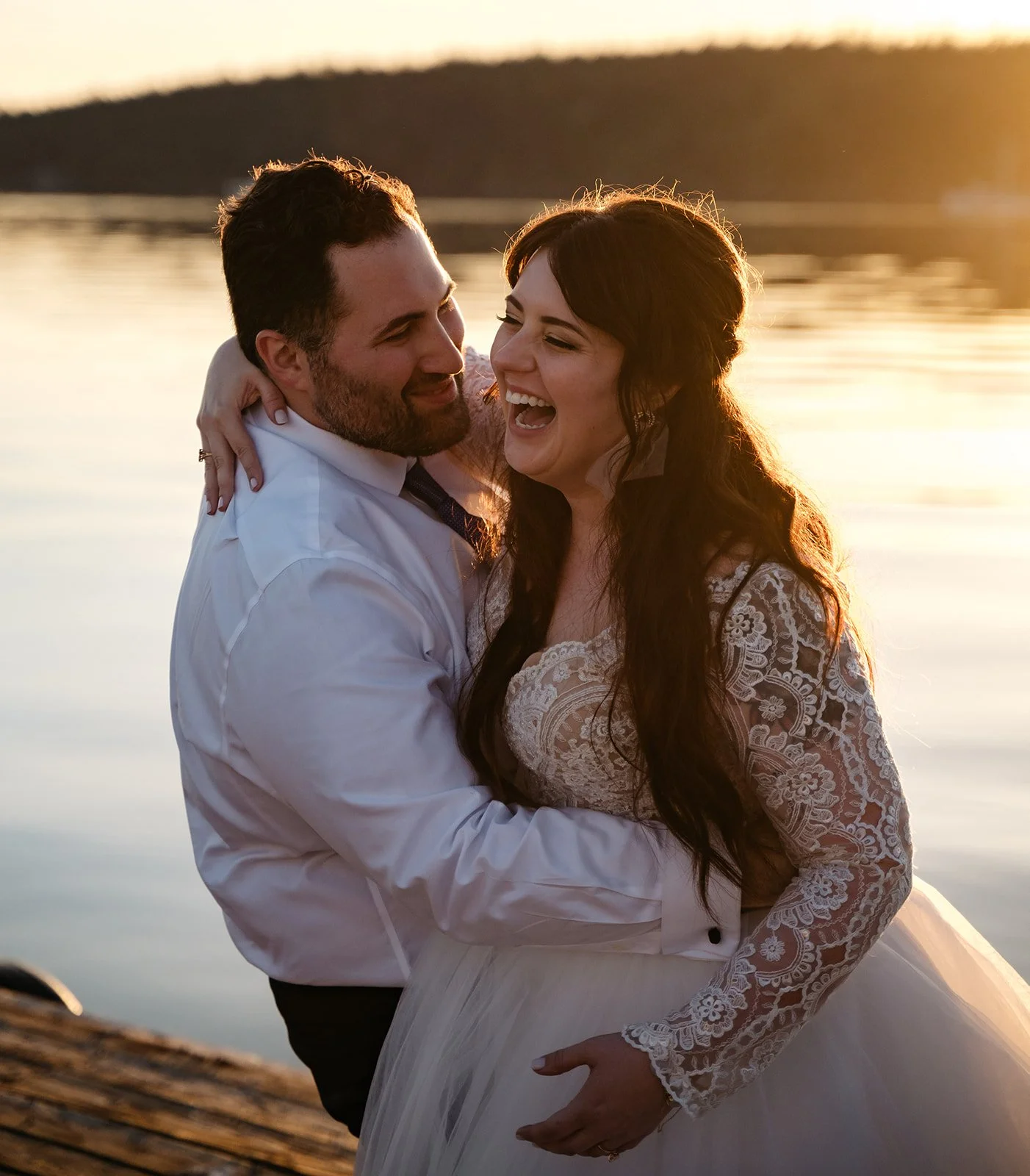 A couple in wedding attire sharing a joyful embrace by a lake at sunset.