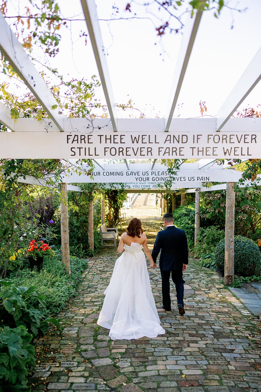 A bride and groom walking hand in hand through a garden pathway, surrounded by greenery and flowers, during a wedding celebration.