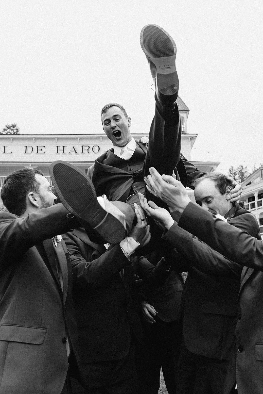 A group of men dressed in tuxedos are celebrating and lifting a man in a suit into the air. The man appears excited and happy, with his mouth open and one leg extended upward. The photograph is in black and white.