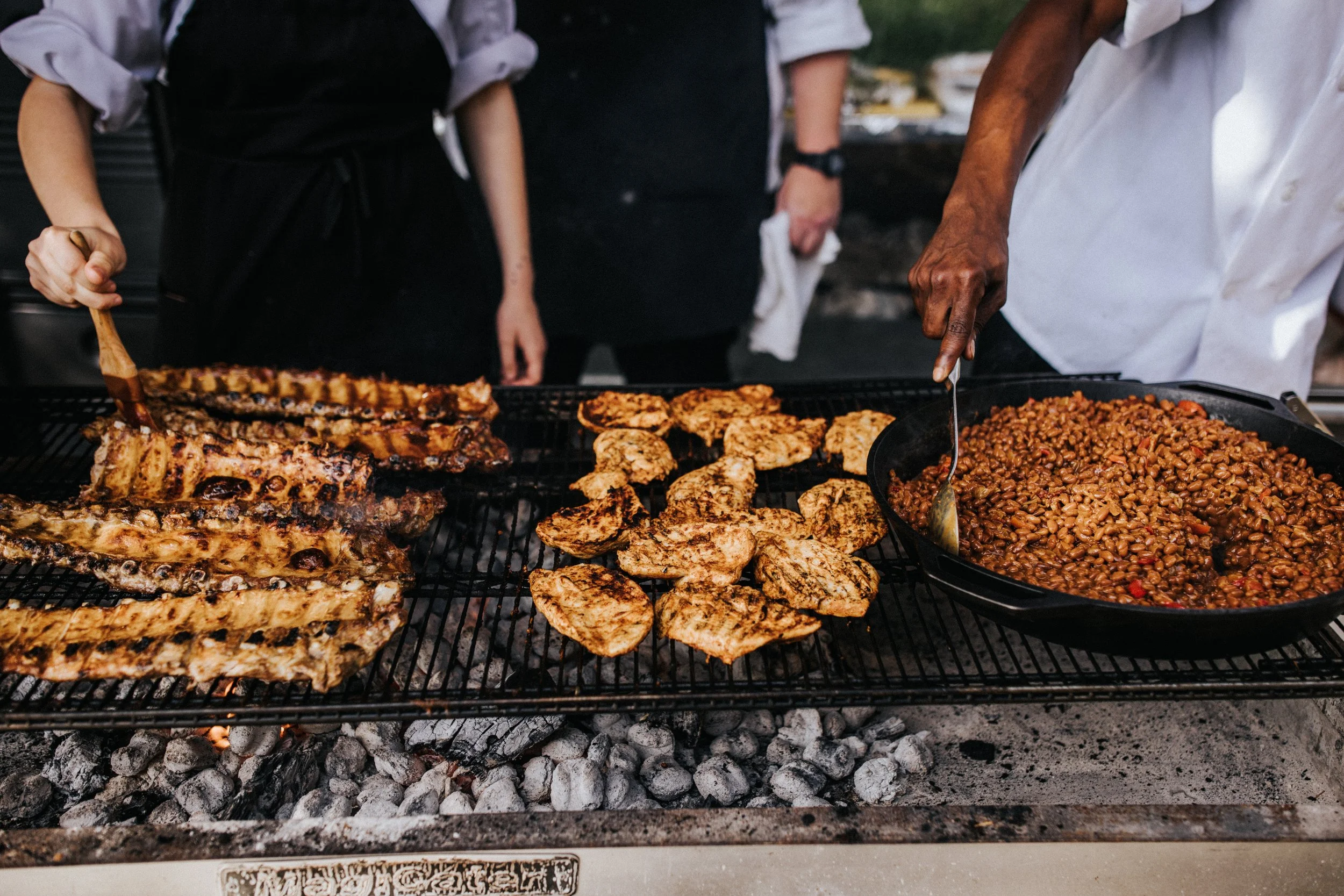 Barbecue grill with ribs, chicken, baked beans, and grilled vegetables, with people cooking and serving food.