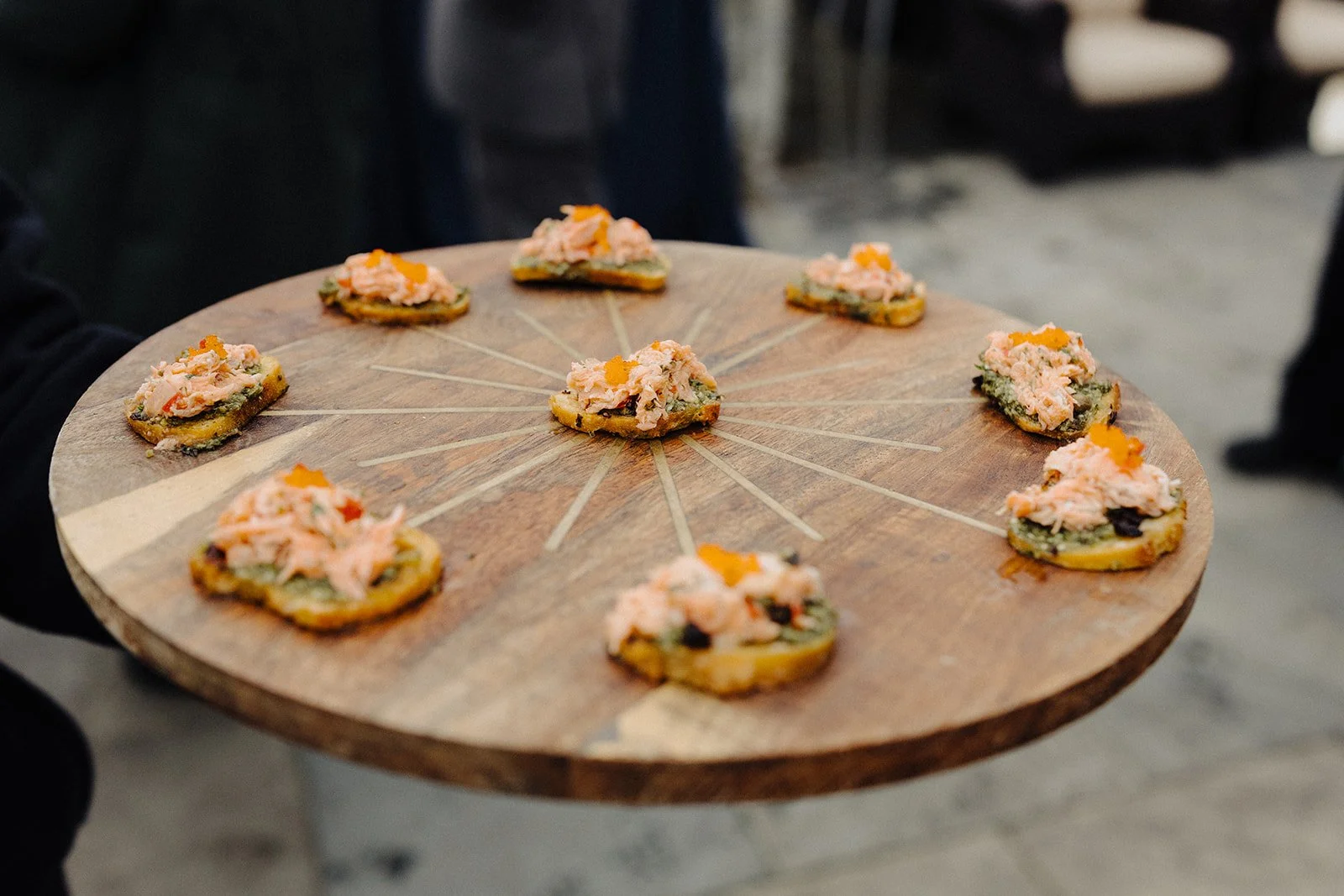 A wooden platter with nine small appetizers topped with pinkish fish salad and orange garnish, arranged in a circle.