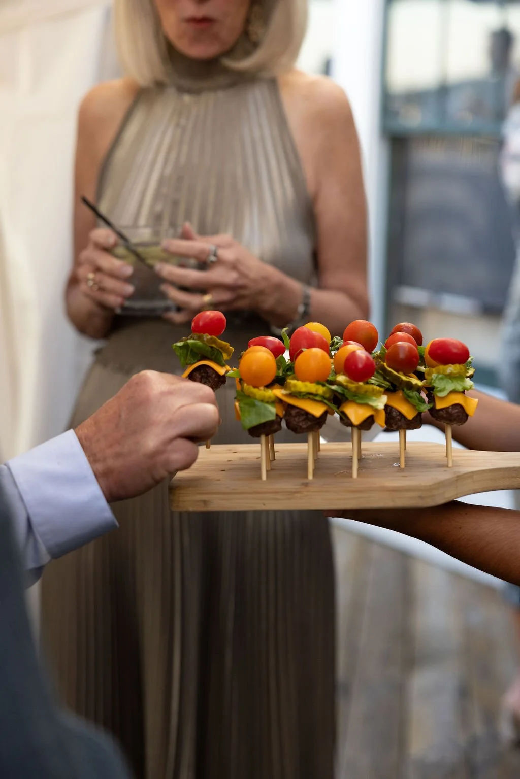 Person holding a wooden tray with mini appetizers topped with cherry tomatoes, lettuce, cheese, and meat, while another person, partially visible, is reaching for one.