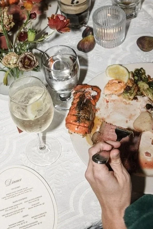 A dinner table with a floral centerpiece, glasses of water and white wine, a plate with lobster, sliced meat, potatoes, and vegetables, a hand holding a piece of meat, and a dinner menu.