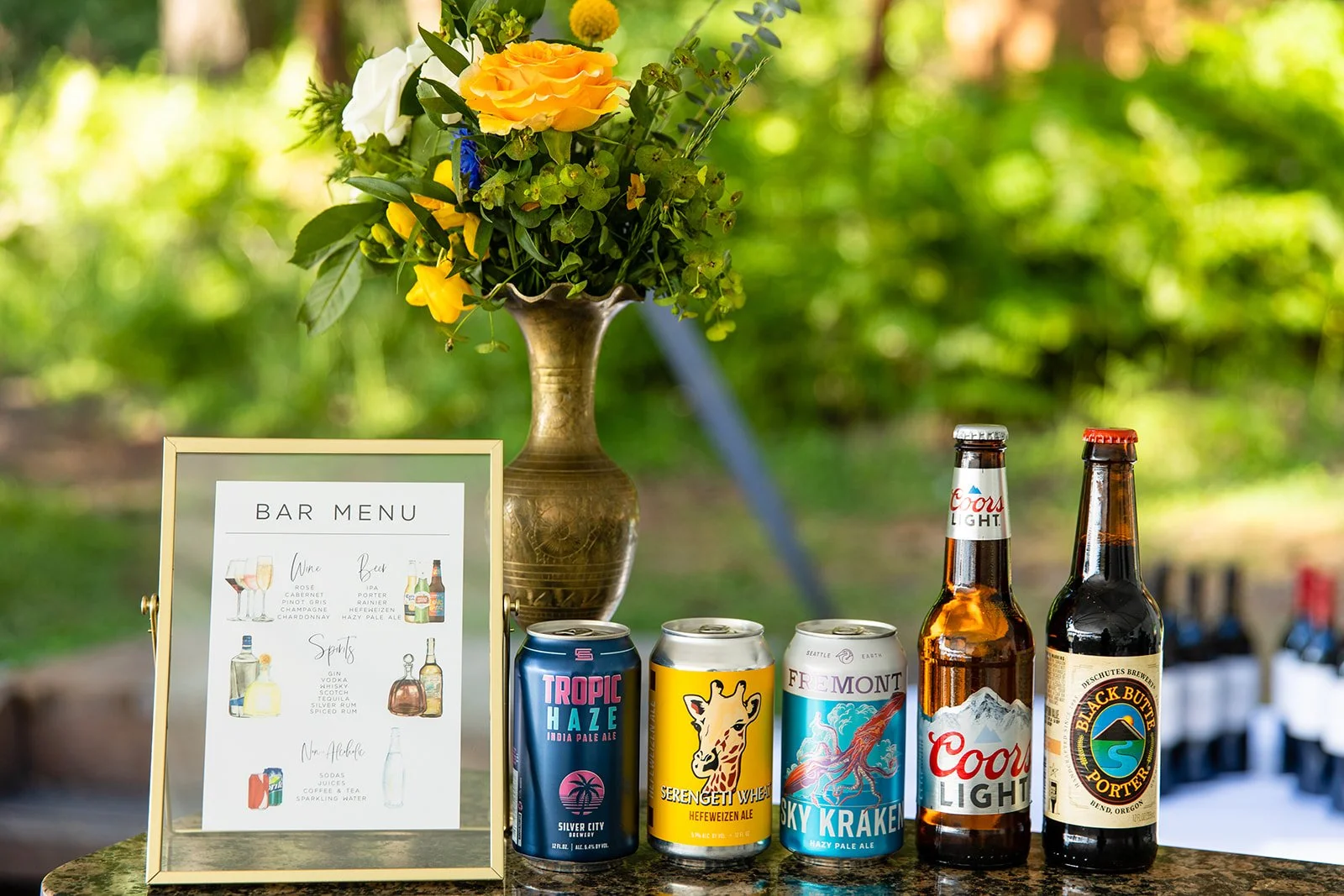 An outdoor bar setup with a floral arrangement in a decorative vase, a bar menu sign, and various beers including Tropic Haze, Serengetti Wheat, Sky Kraken, Coors Light, and Black Butte Porter on a table.