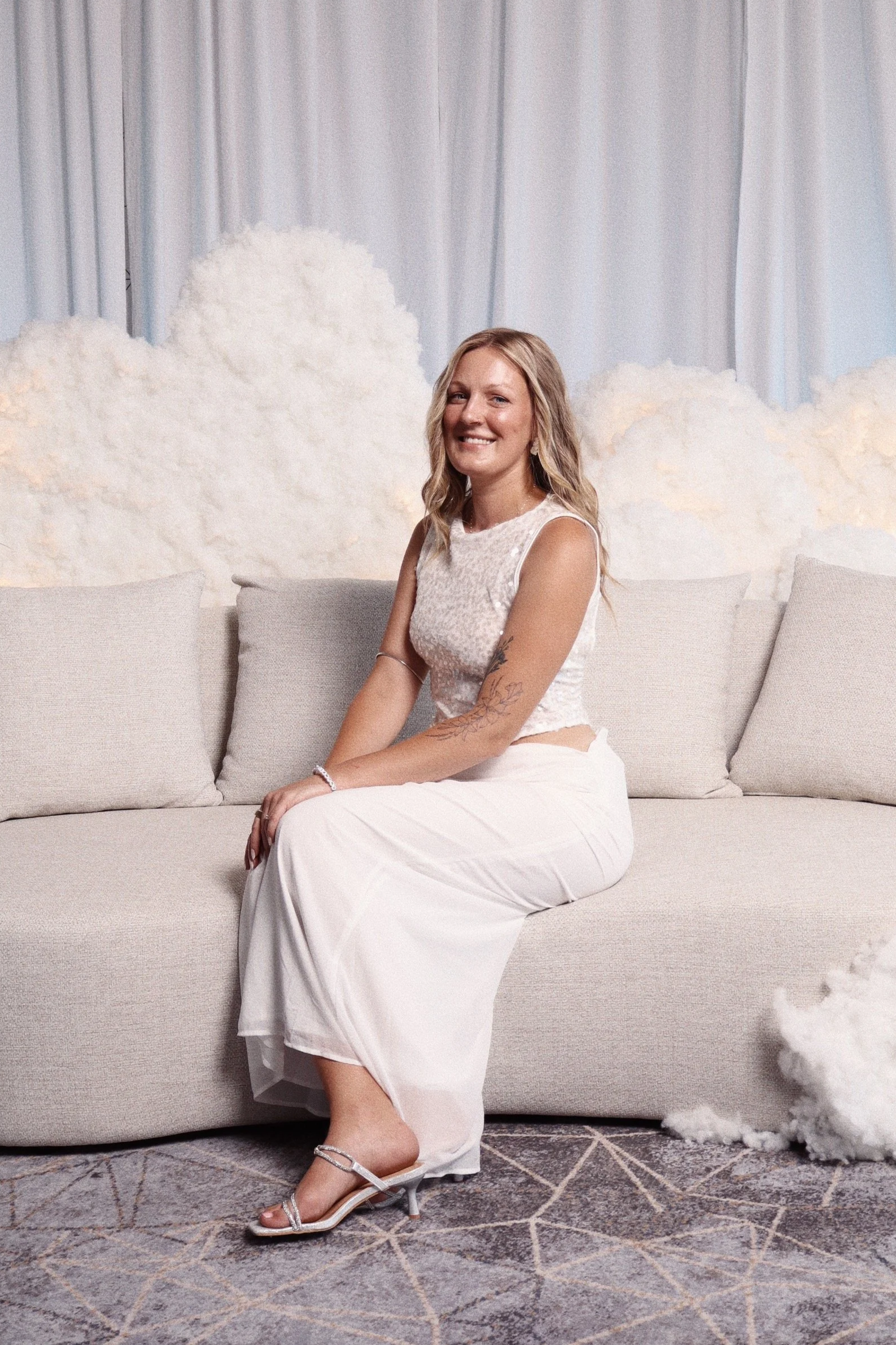 A woman sitting on a beige sofa with cloud decorations behind her, smiling at the camera, dressed in a white sleeveless top, white skirt, and silver high heels.