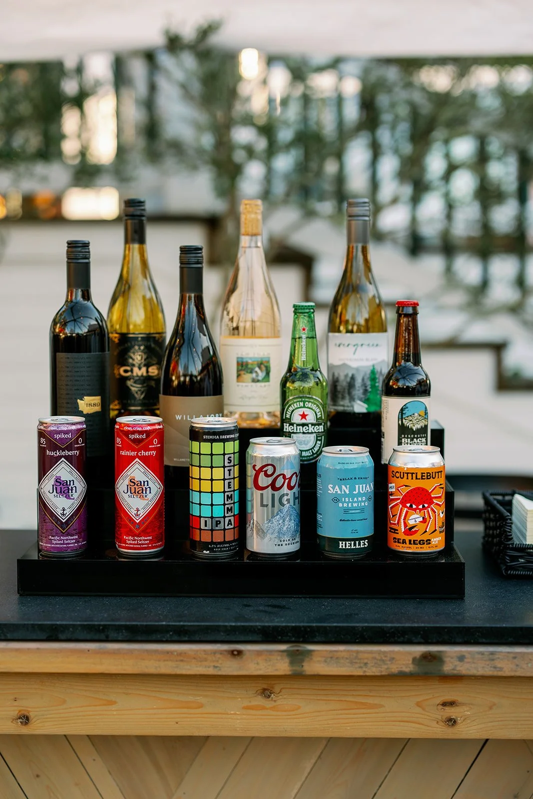 A selection of bottled beer and canned drinks on a wooden bar countertop outdoors, with trees and a white railing in the background.
