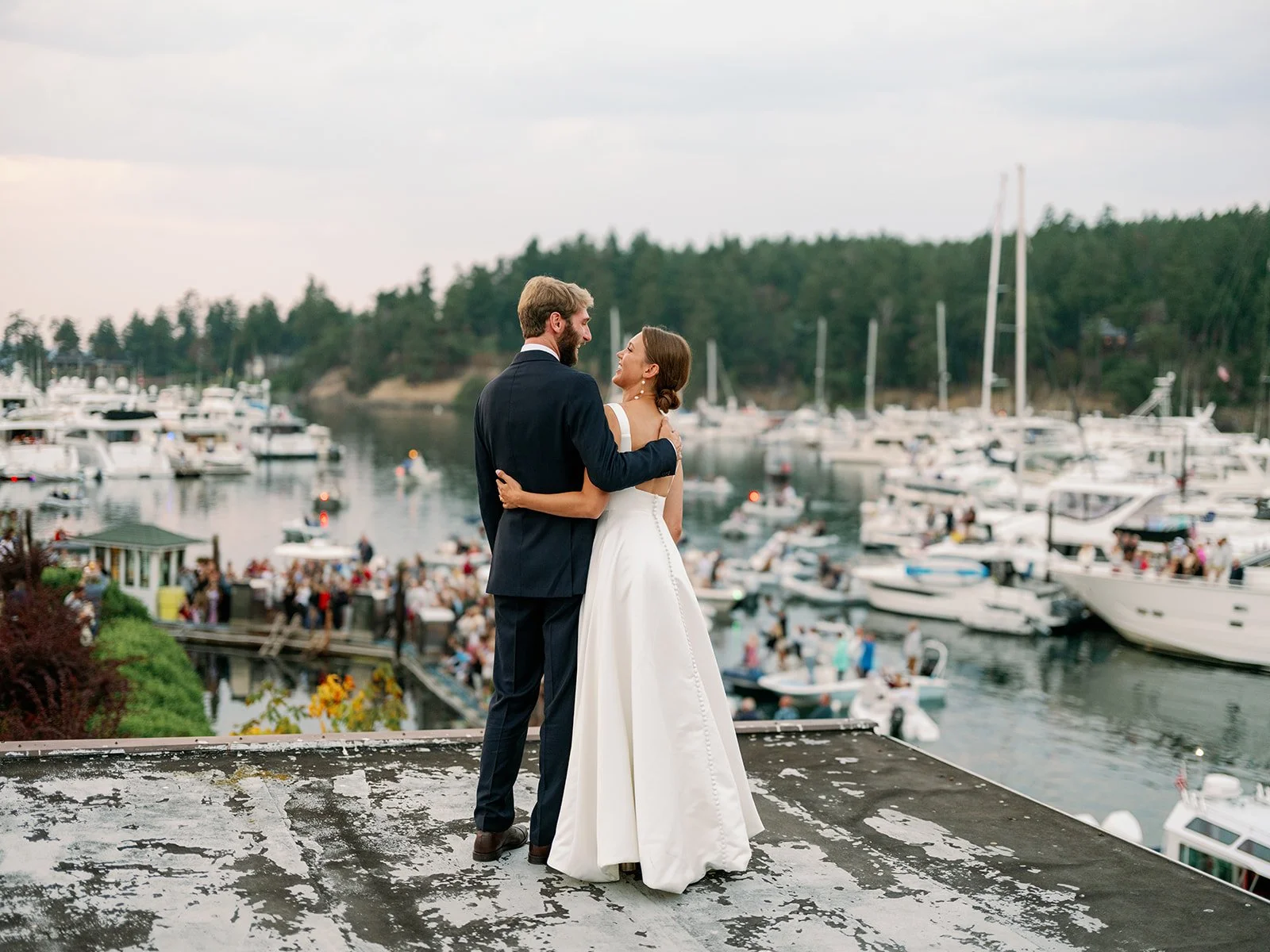 A newlywed couple stands on a rooftop overlooking a marina with boats, holding each other and smiling, with a backdrop of water and greenery.