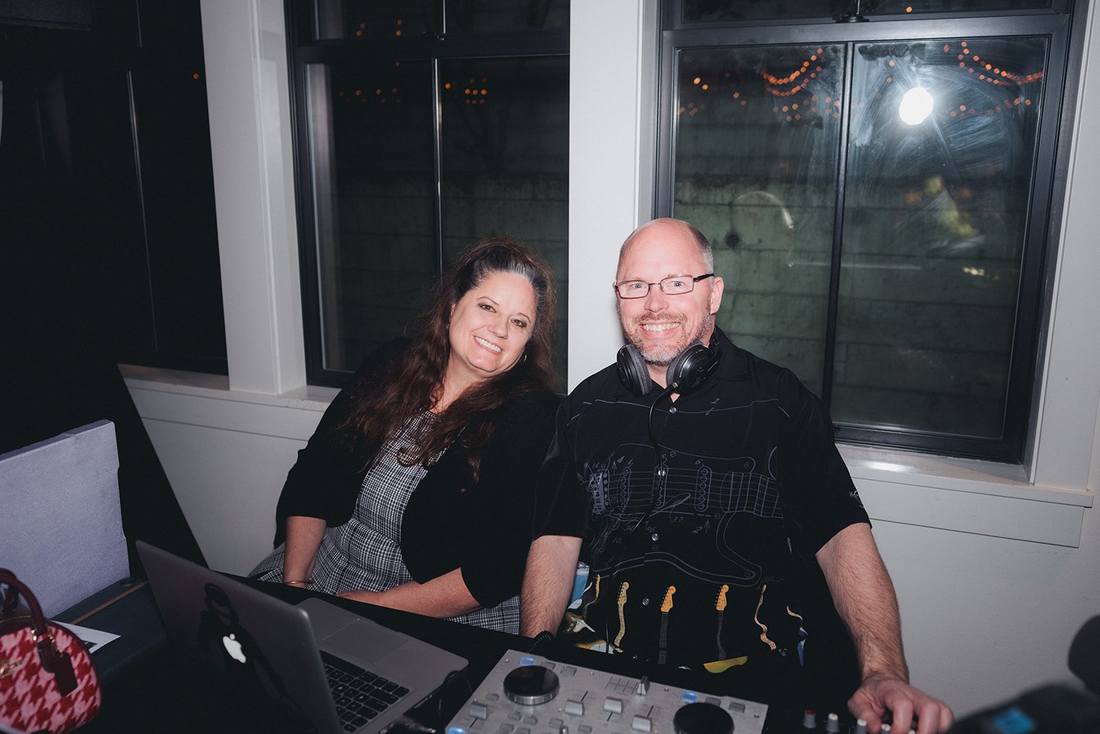 A smiling woman with dark curly hair and a man with glasses and a beard wearing headphones, sitting at a table with DJ equipment and a laptop, in front of a window at night.