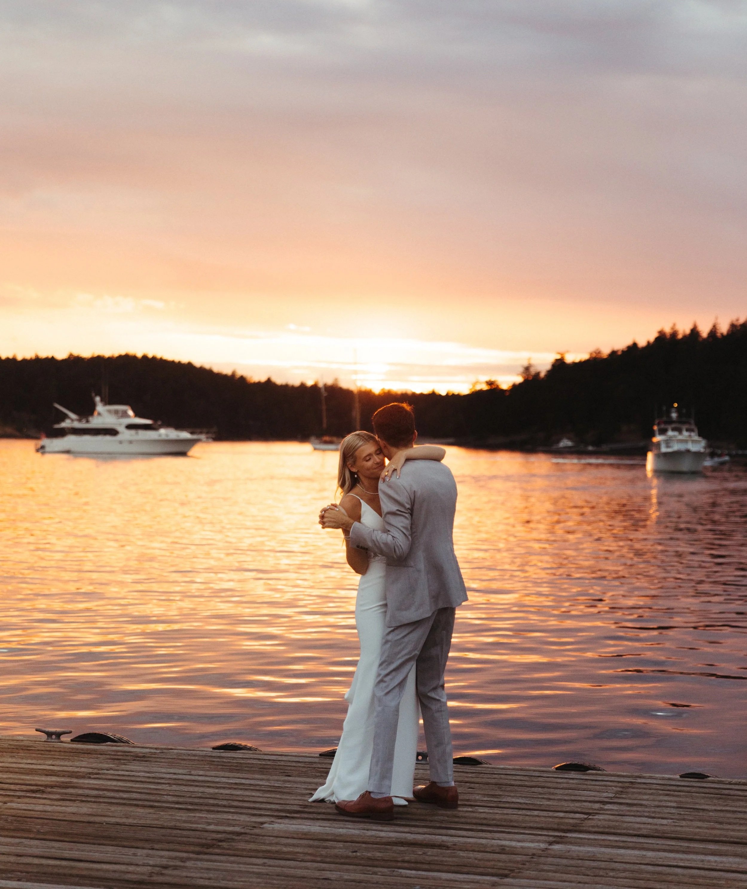 A couple in wedding attire dancing on a wooden dock by the water during sunset, with boats anchored in the background and a colorful sky.