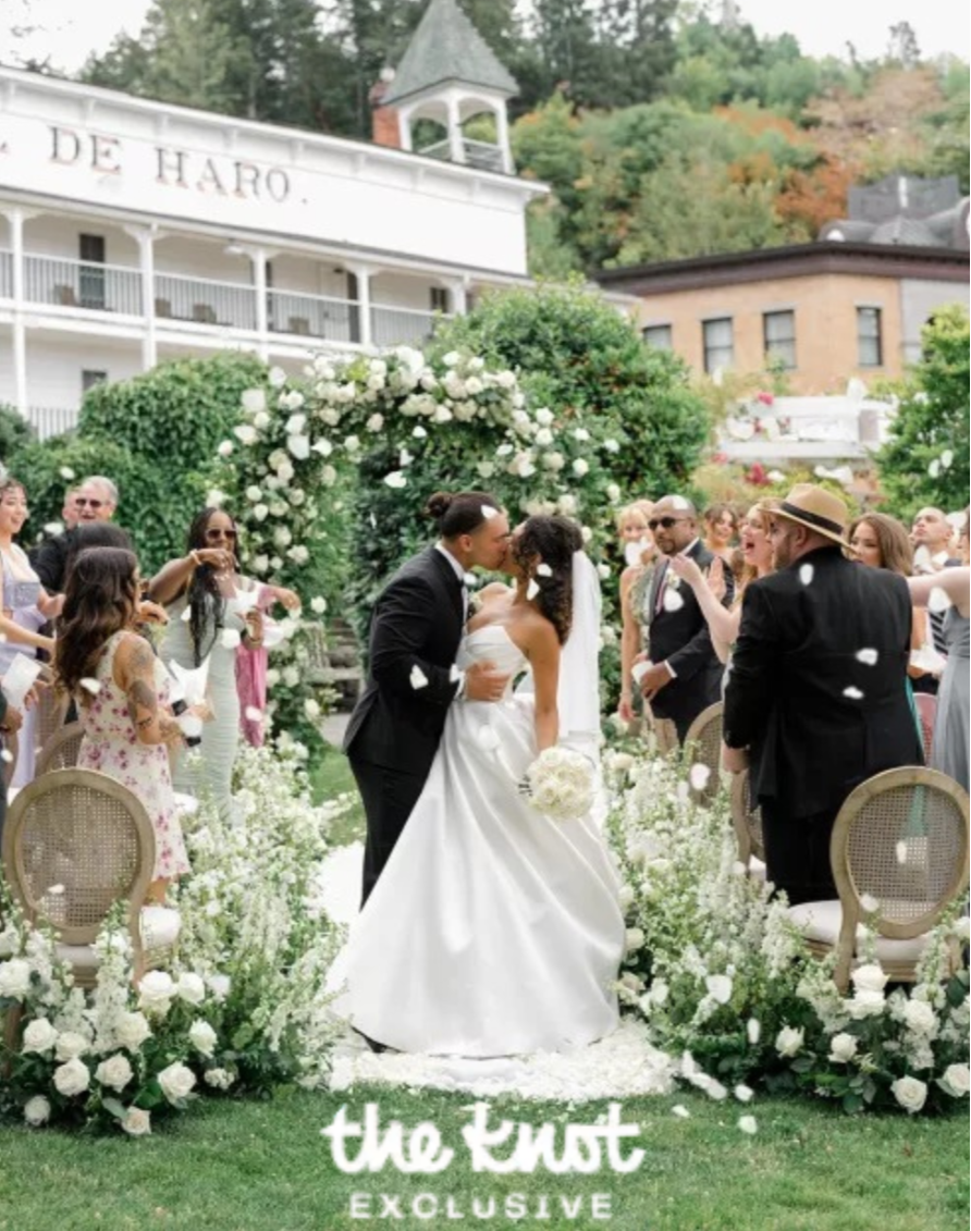 A bride and groom sharing a kiss during their outdoor wedding ceremony, surrounded by guests and decorated with white flowers and greenery.