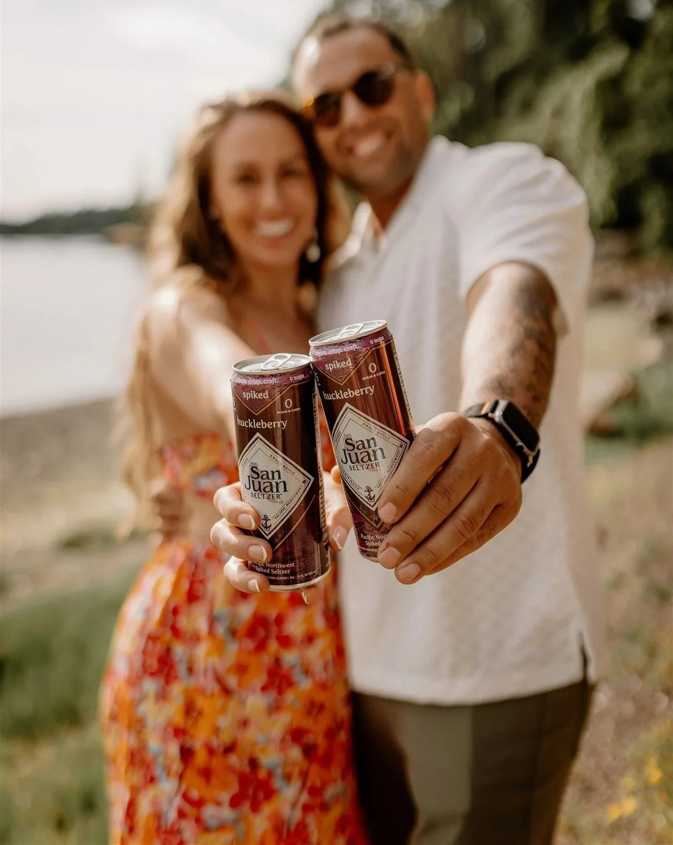 A smiling couple outdoors holding cans of San Juan Seltzer close to the camera, with a blurred background of water and trees.