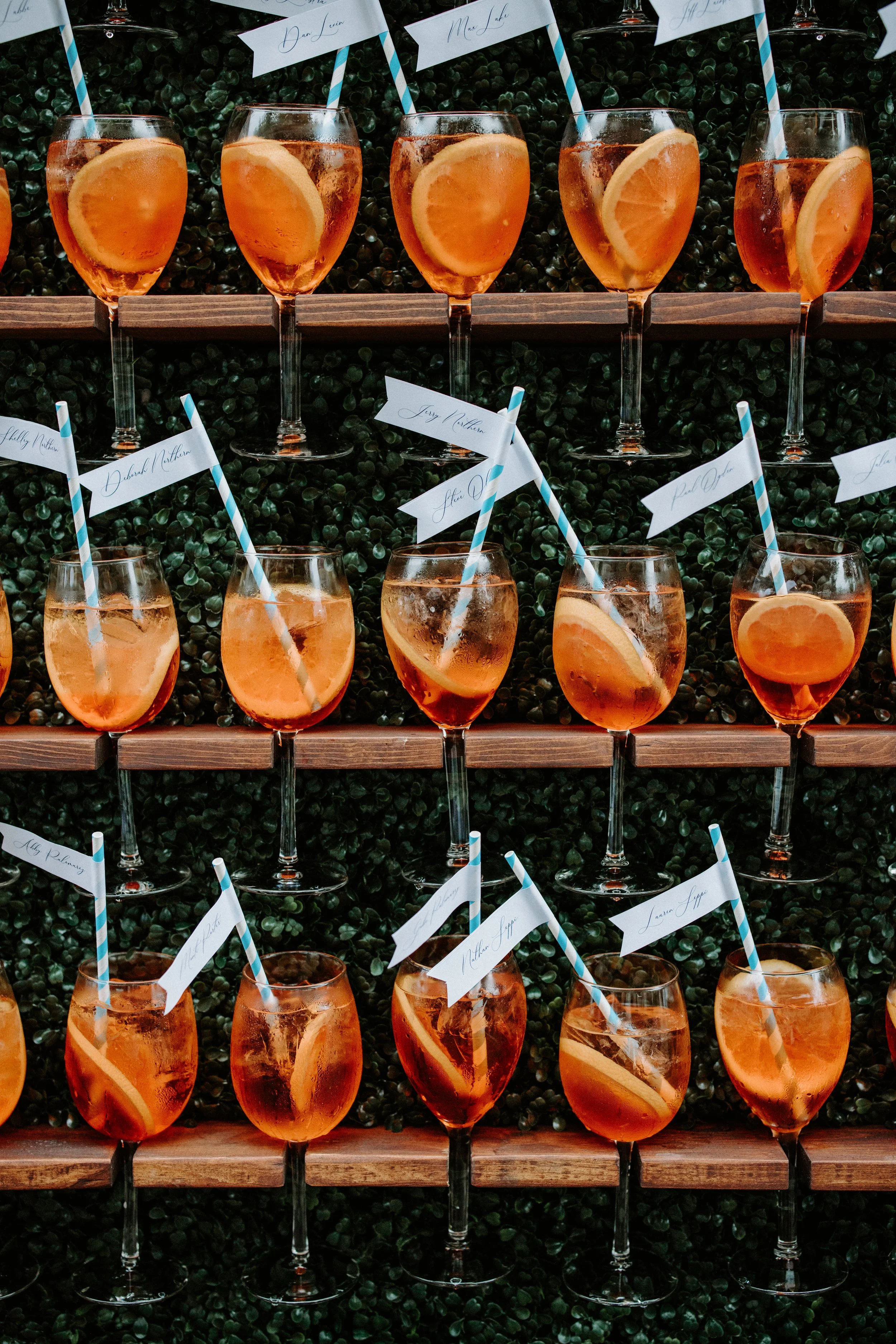 Multiple glasses of peach-colored drinks with lemon slices and paper flags with cursive writing, displayed on a wooden and green leafy backdrop.