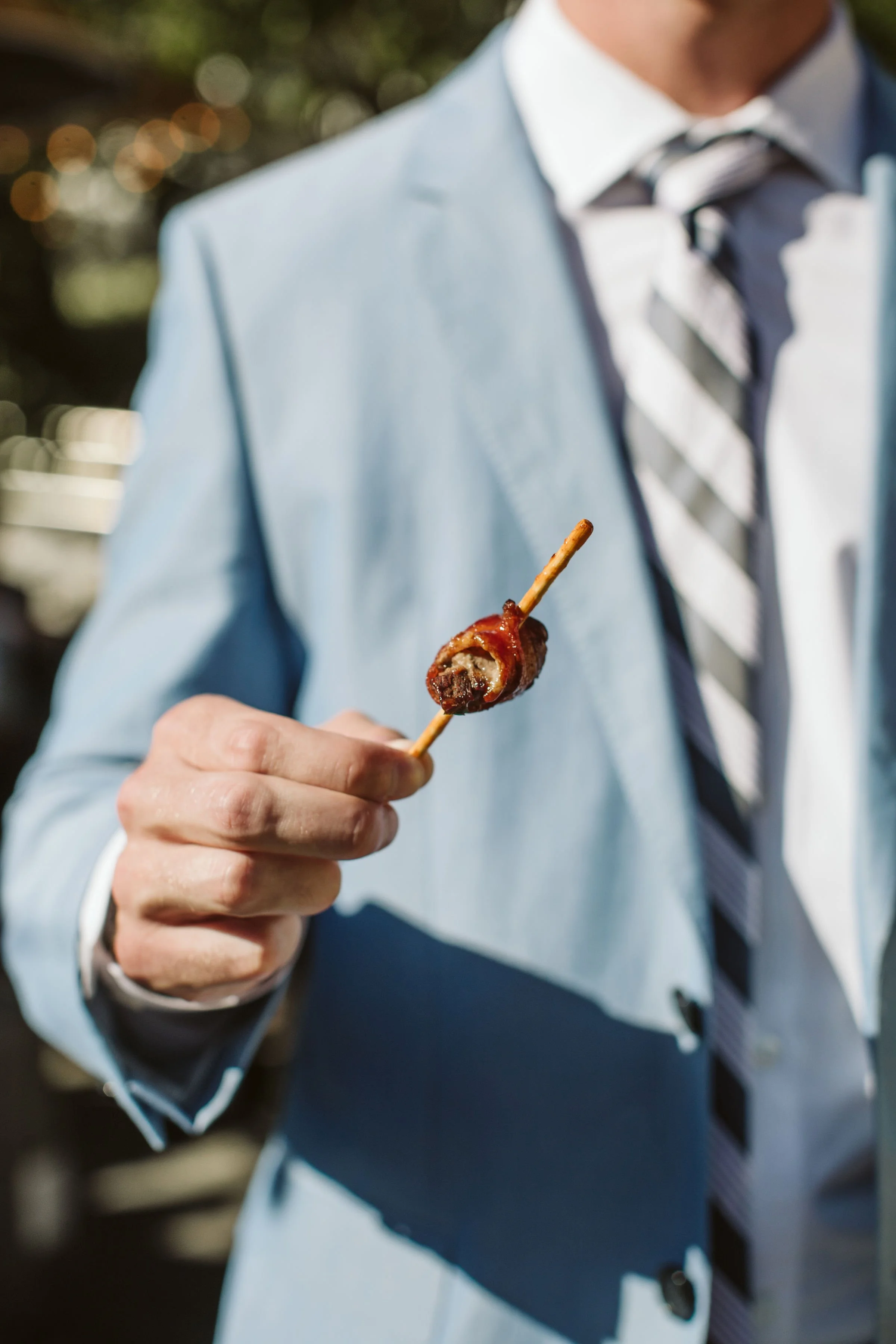 A man in a blue suit and striped tie holding a bacon-wrapped scallop on a toothpick.