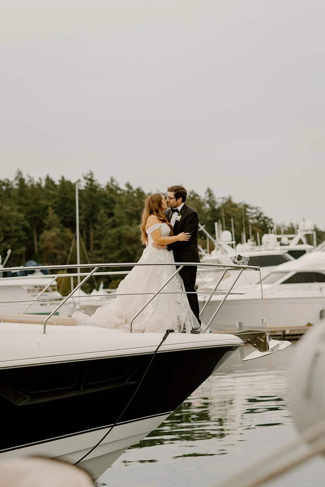 A newlywed couple sharing a kiss on a yacht docked at a marina, with boats and trees in the background.