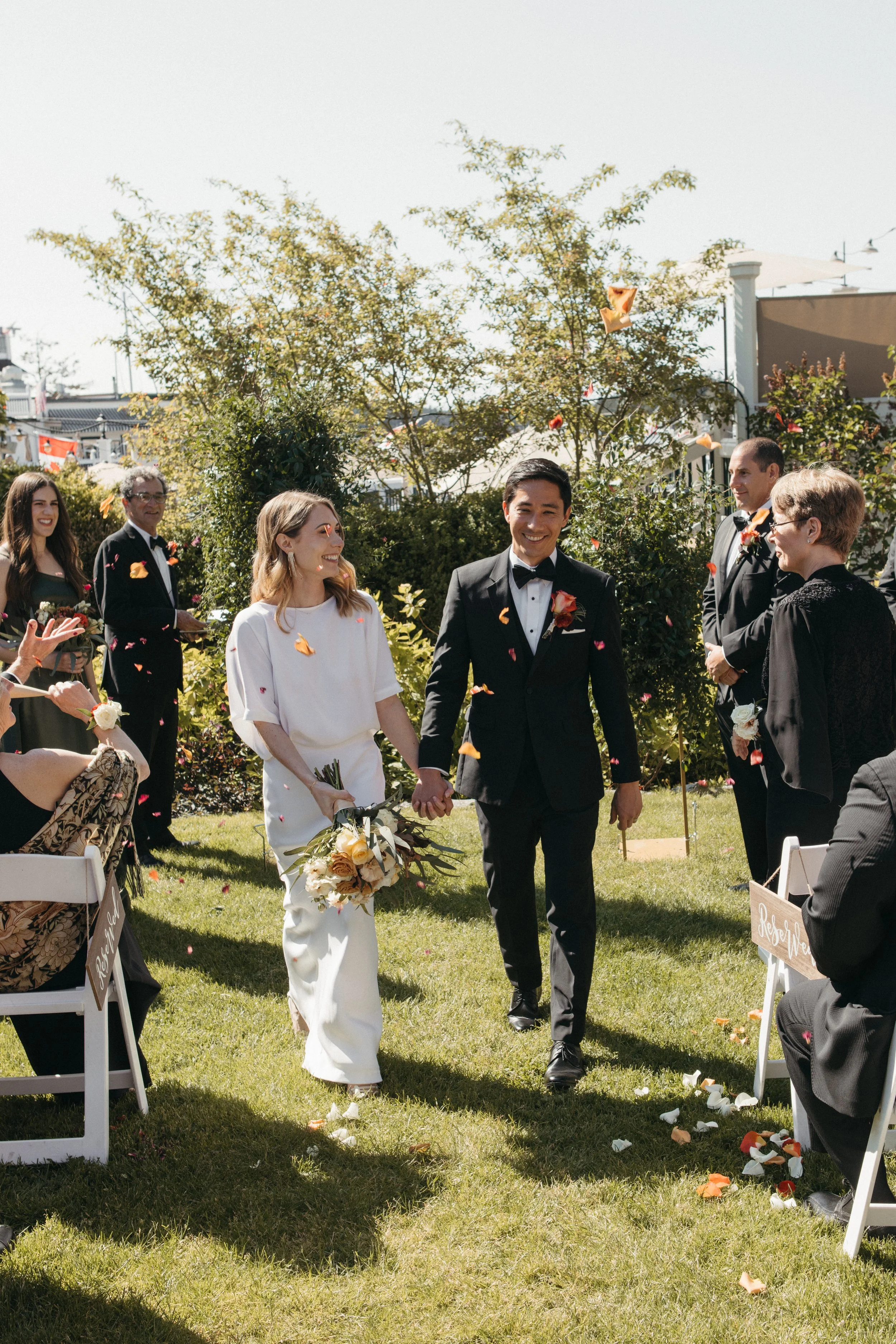 Couple holding hands, walking down aisle at outdoor wedding, surrounded by guests, flower petals in the air, greenery, sunny weather.