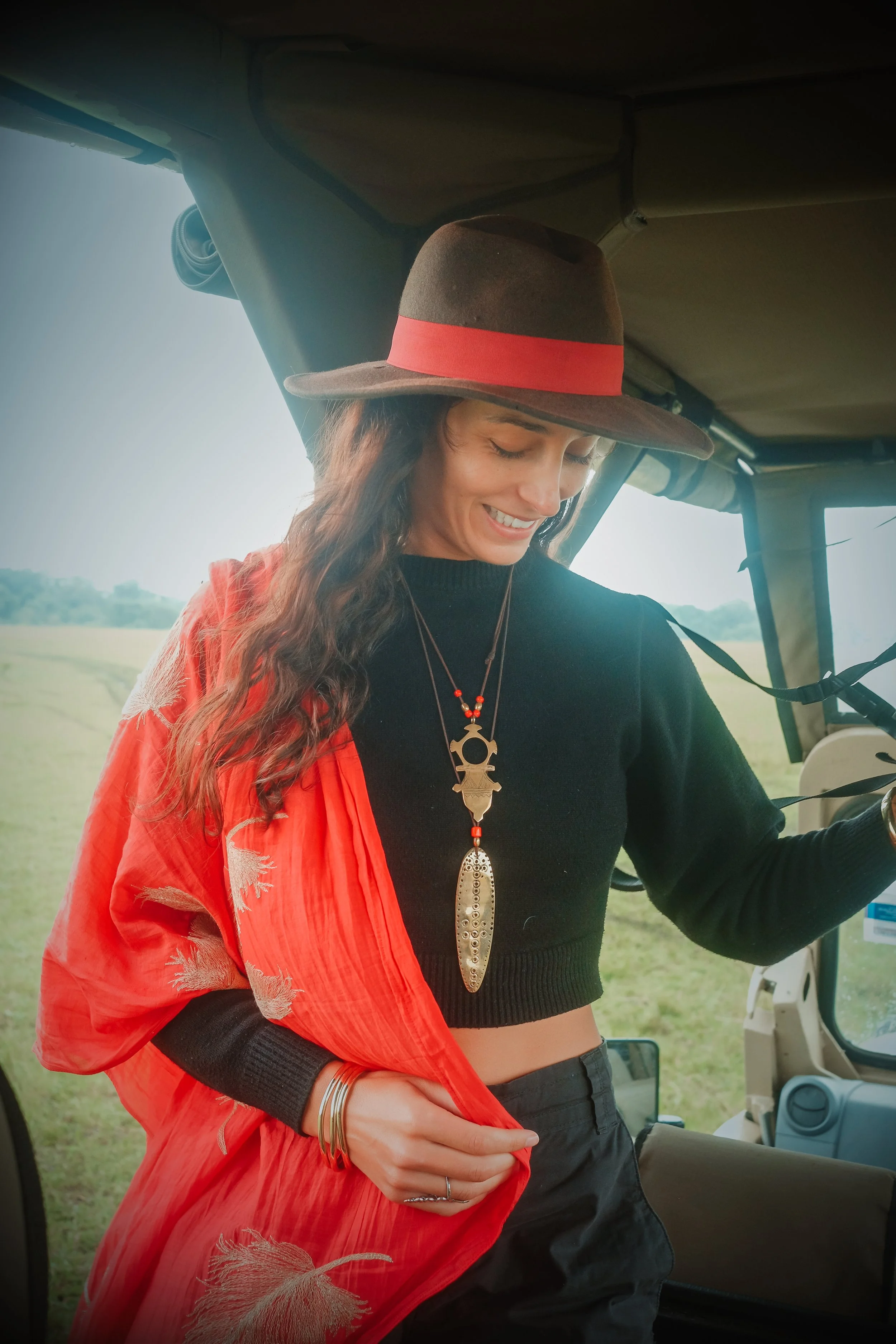 A woman with long wavy hair wearing a wide-brimmed hat with a red band, a black crop top, and layered necklaces, standing inside a vehicle with a grassy outdoor background.