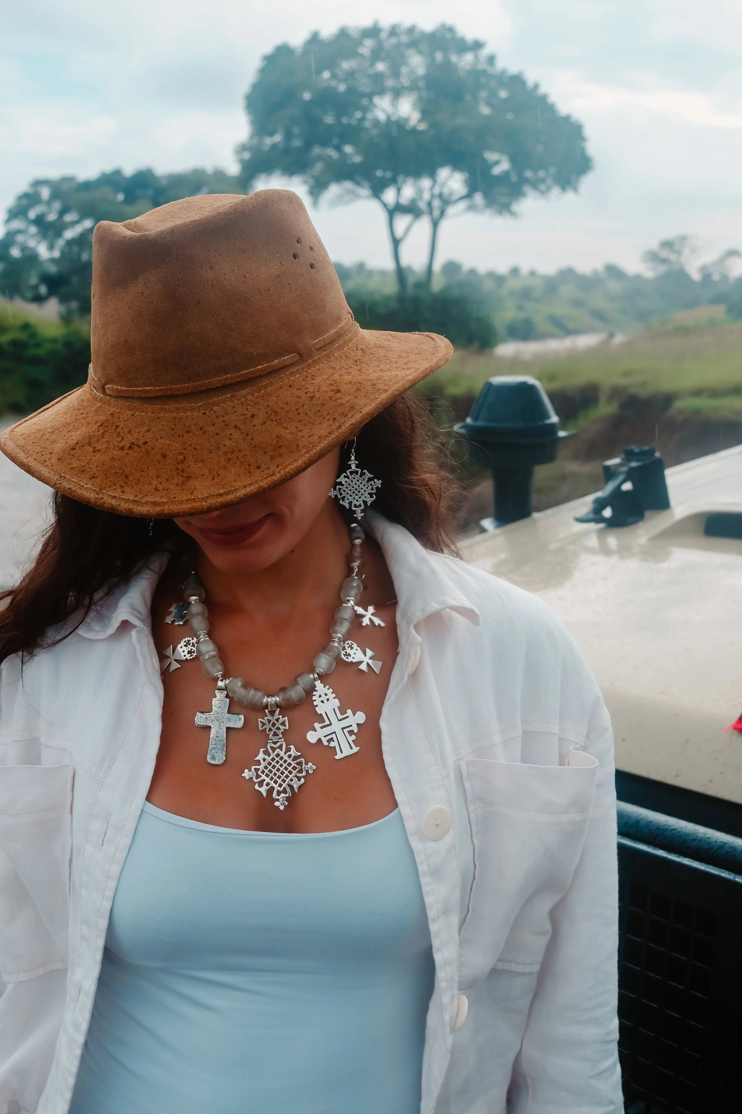 A woman wearing a brown hat, a white open shirt, and a large silver necklace with crosses and other designs, standing outdoors near a boat or vehicle with a landscape of trees and water in the background.