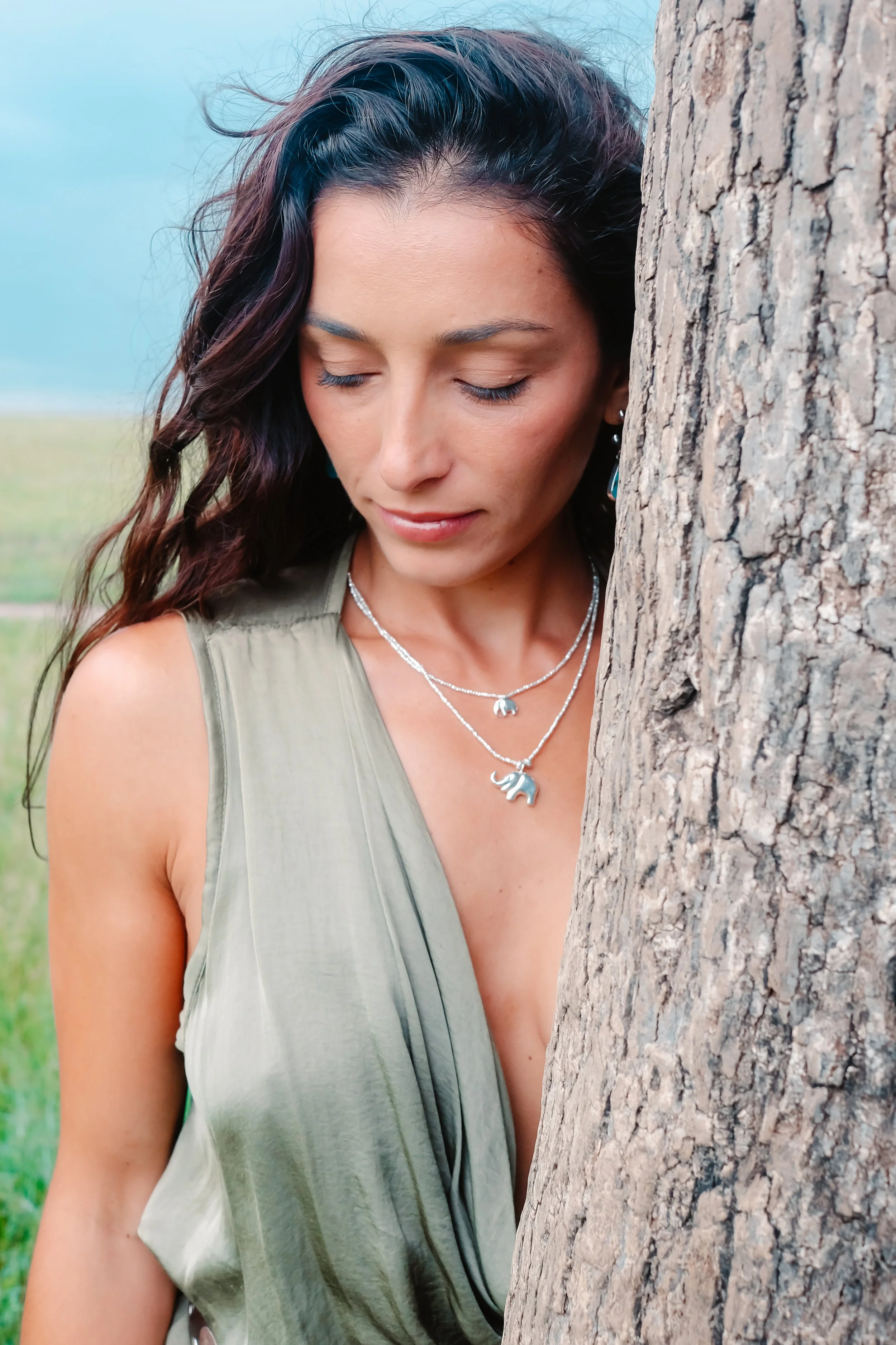 A woman with dark wavy hair and light skin, eyes closed, leaning against a tree outdoors, wearing a sleeveless olive-green top and silver necklaces with animal pendants.