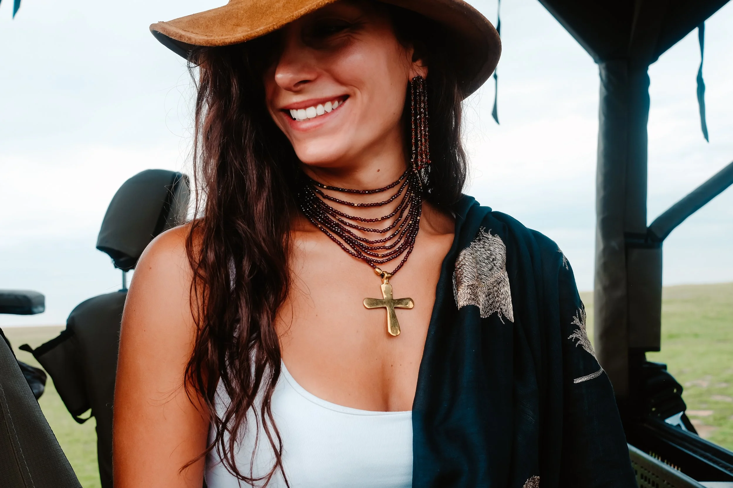 A smiling woman wearing a wide-brimmed hat, layered necklaces including a large gold cross, and a black garment with embroidery, standing outdoors.