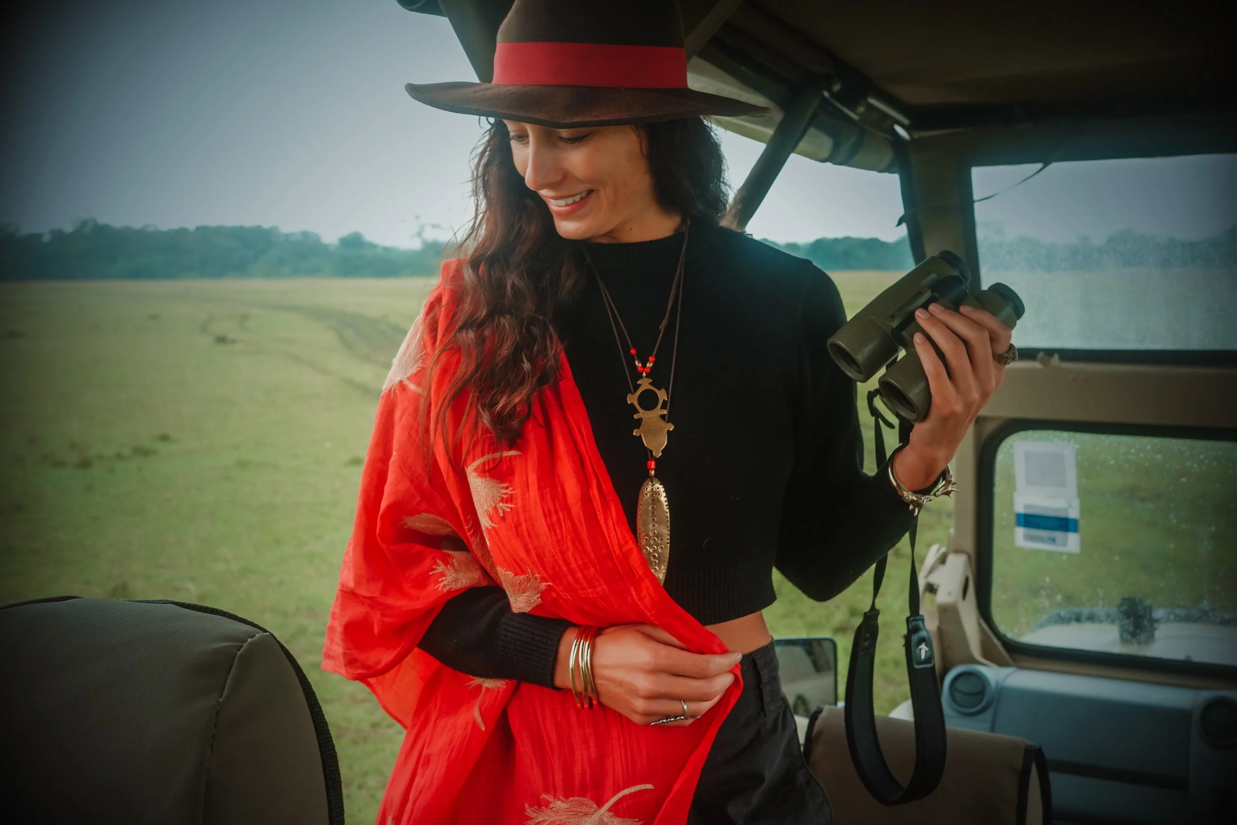 Woman in a large wide-brimmed hat, black top, and red shawl, holding binoculars and smiling inside a safari vehicle with a grassy plain background.