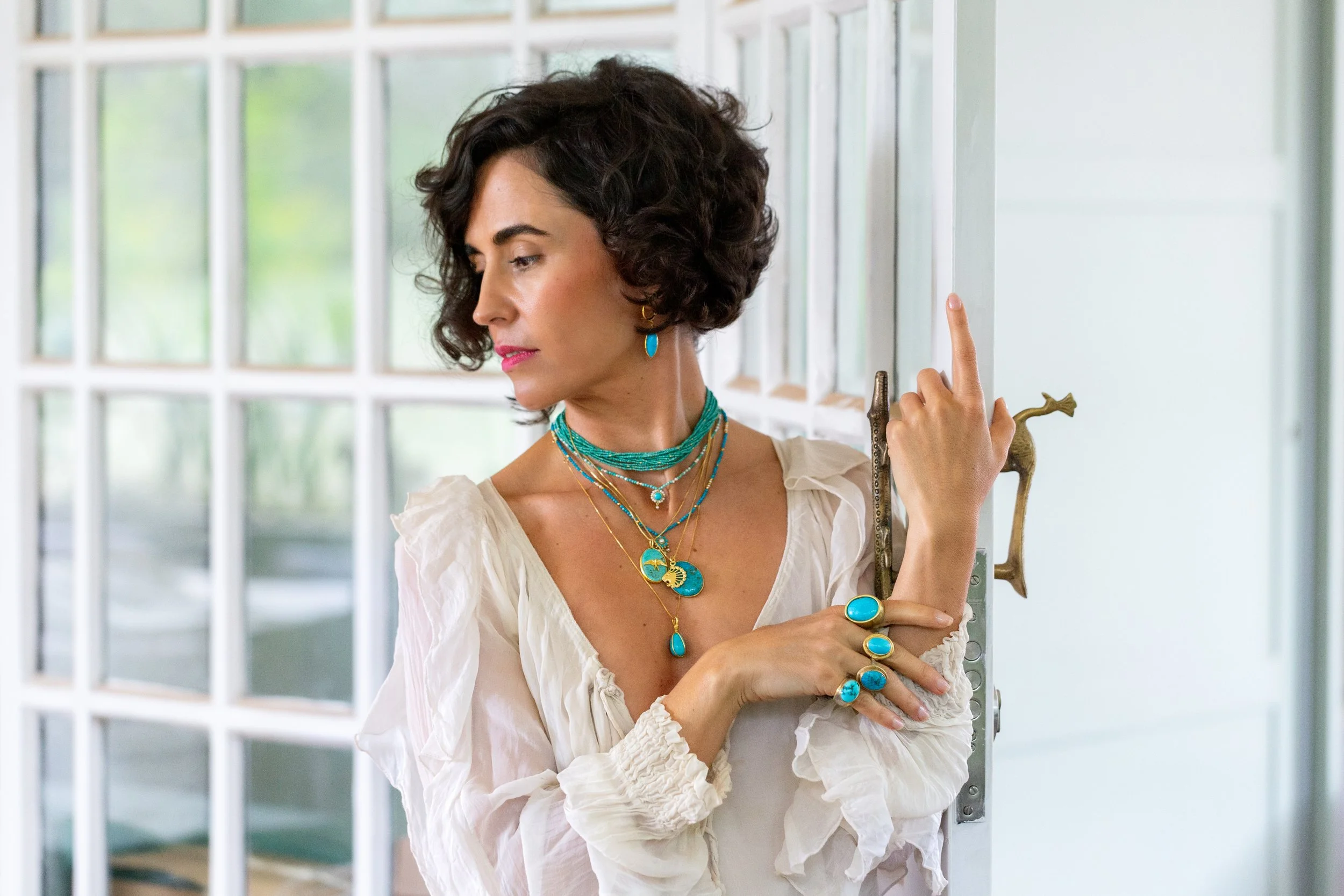A woman with short curly dark hair wearing multiple turquoise jewelry pieces, a cream-colored blouse, and standing by a door with a brass handle.