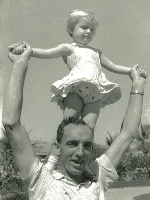 A man with a woman on his shoulders, holding her hands, outdoors with trees and sky visible