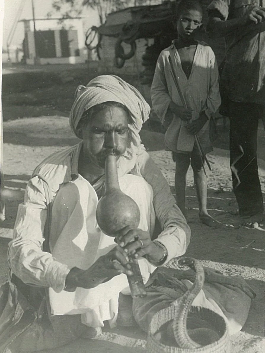A man with a turban blowing into a horn while sitting on the ground, with a snake coiled nearby; two children standing behind him in an outdoor setting.