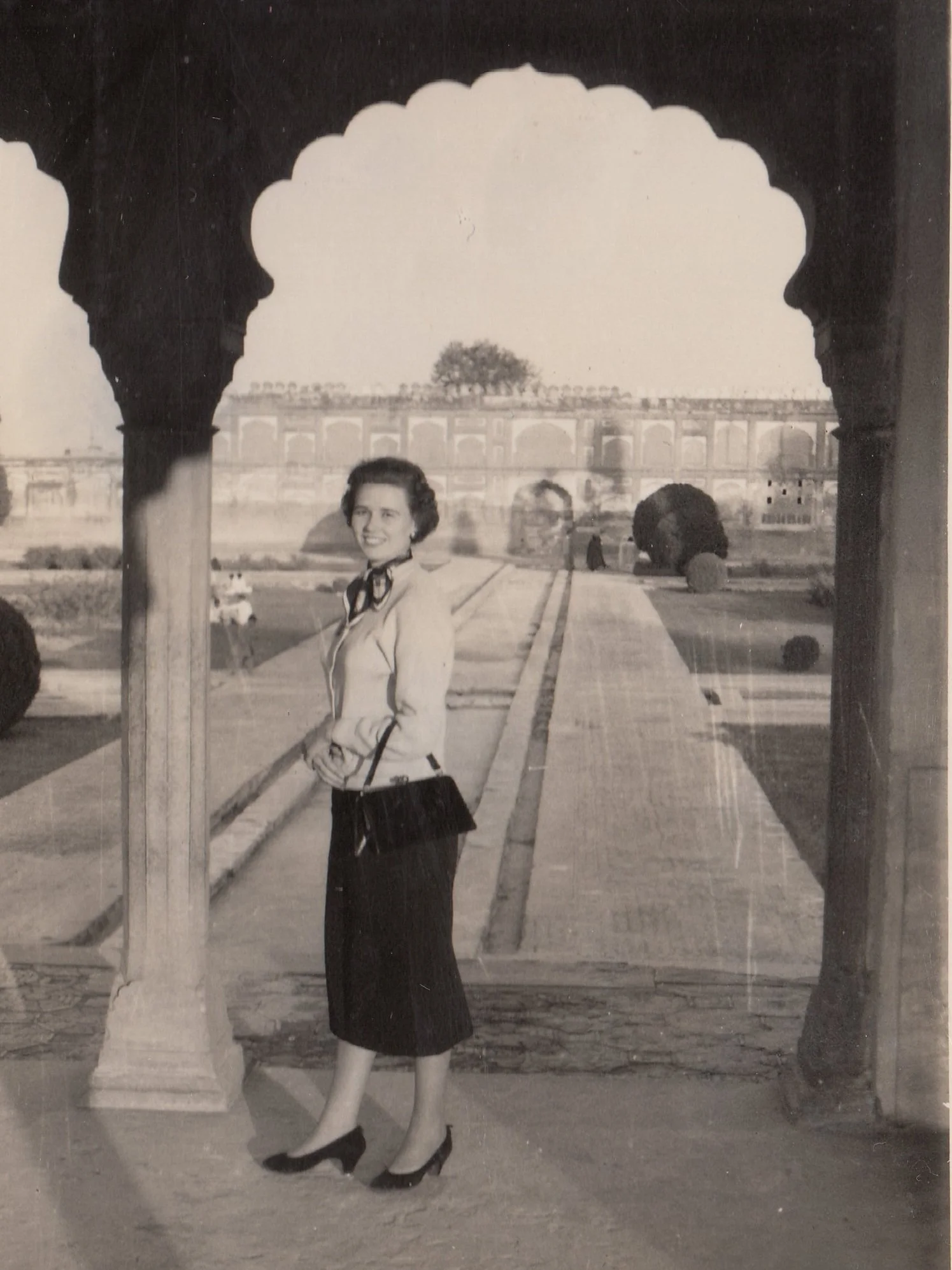 Women standing under an ornate archway, smiling, with a historic building and landscaped garden in the background.