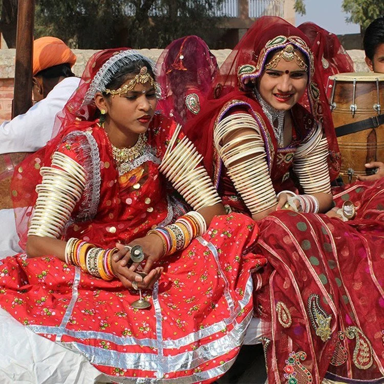 Two women dressed in traditional Indian wedding attire, wearing vibrant red and gold outfits with elaborate jewelry and bangles, seated together outdoors.