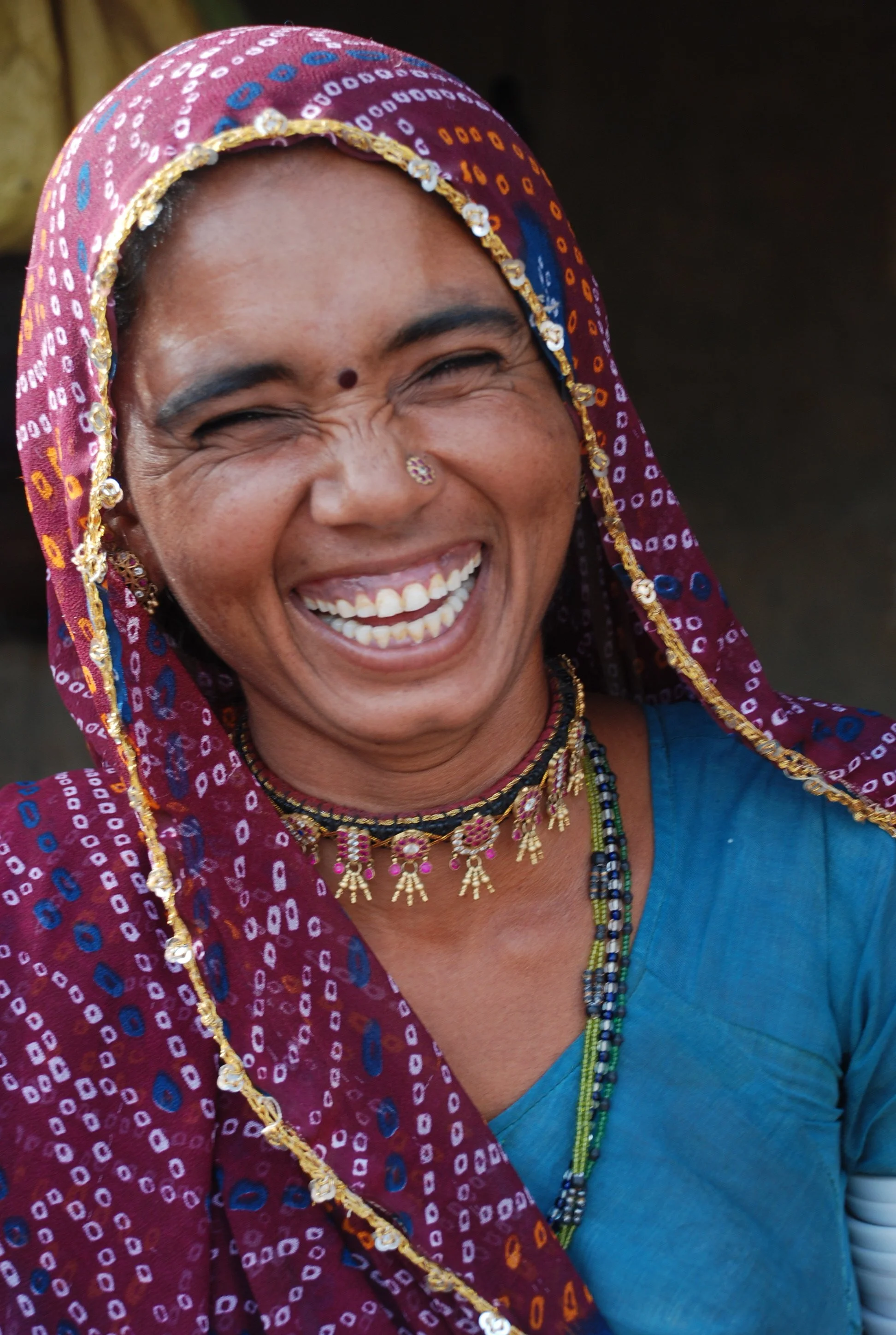 A woman with brown skin is laughing happily, wearing a colorful traditional headscarf and jewelry, with a blue outfit and a blurred dark background.