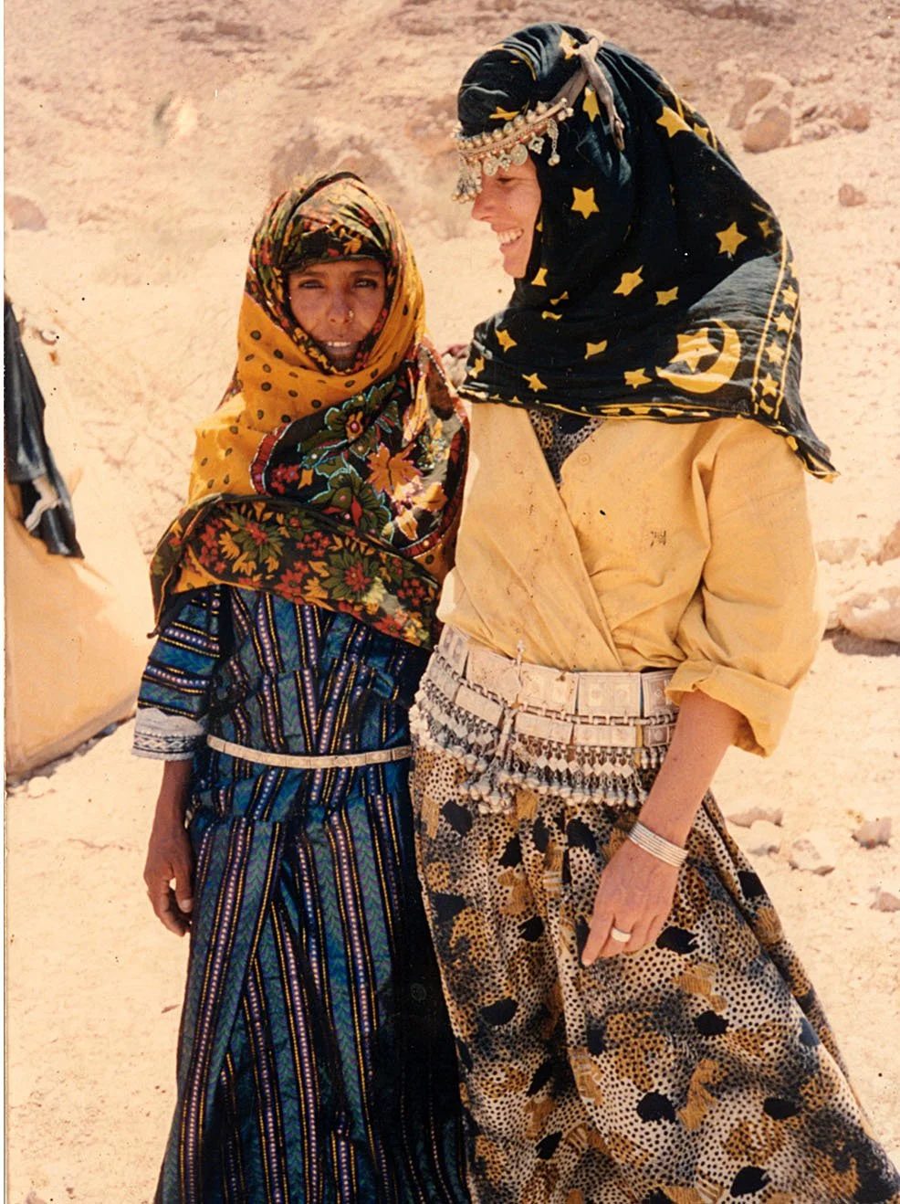 Two women in colorful, patterned clothing and headscarves, smiling together in a desert environment.