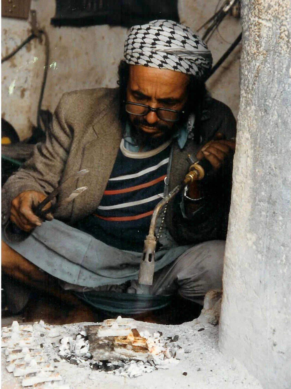 A man in a striped shirt, blazer, and headscarf welds metal pipe near a small fire on the ground indoors.