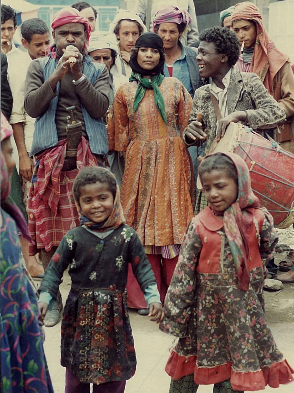 A group of people, including two children in the foreground, are gathered outdoors. The children are smiling, and the group appears to be engaged in a communal event or gathering, with some individuals playing musical instruments.