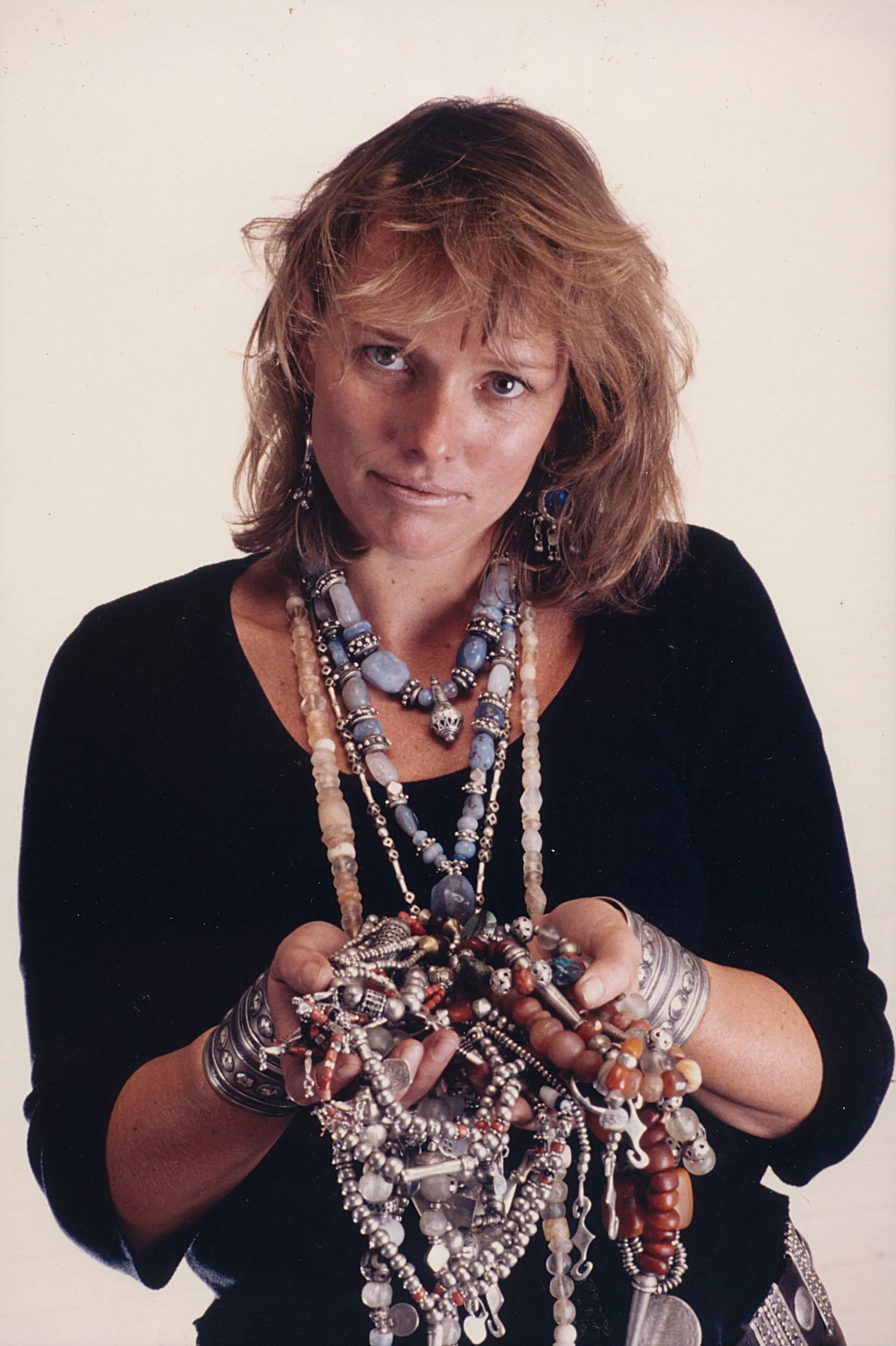 Woman with wavy, shoulder-length hair holding a large collection of jewelry, wearing multiple bracelets, necklaces, and earrings, against a plain background.
