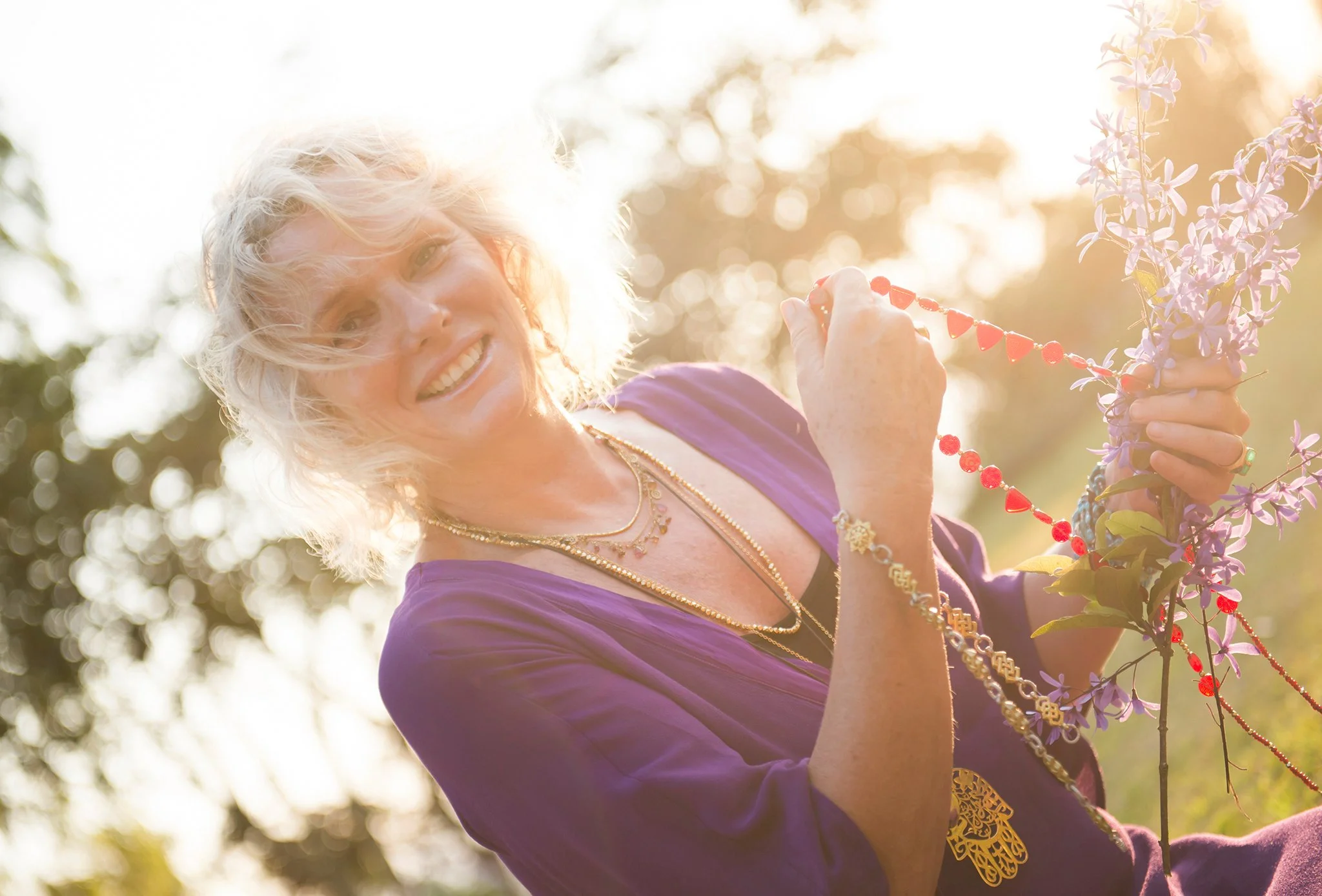A smiling elderly woman with curly blonde hair wearing a purple dress outdoors during golden hour, holding a branch of purple flowers and a string of red beads.