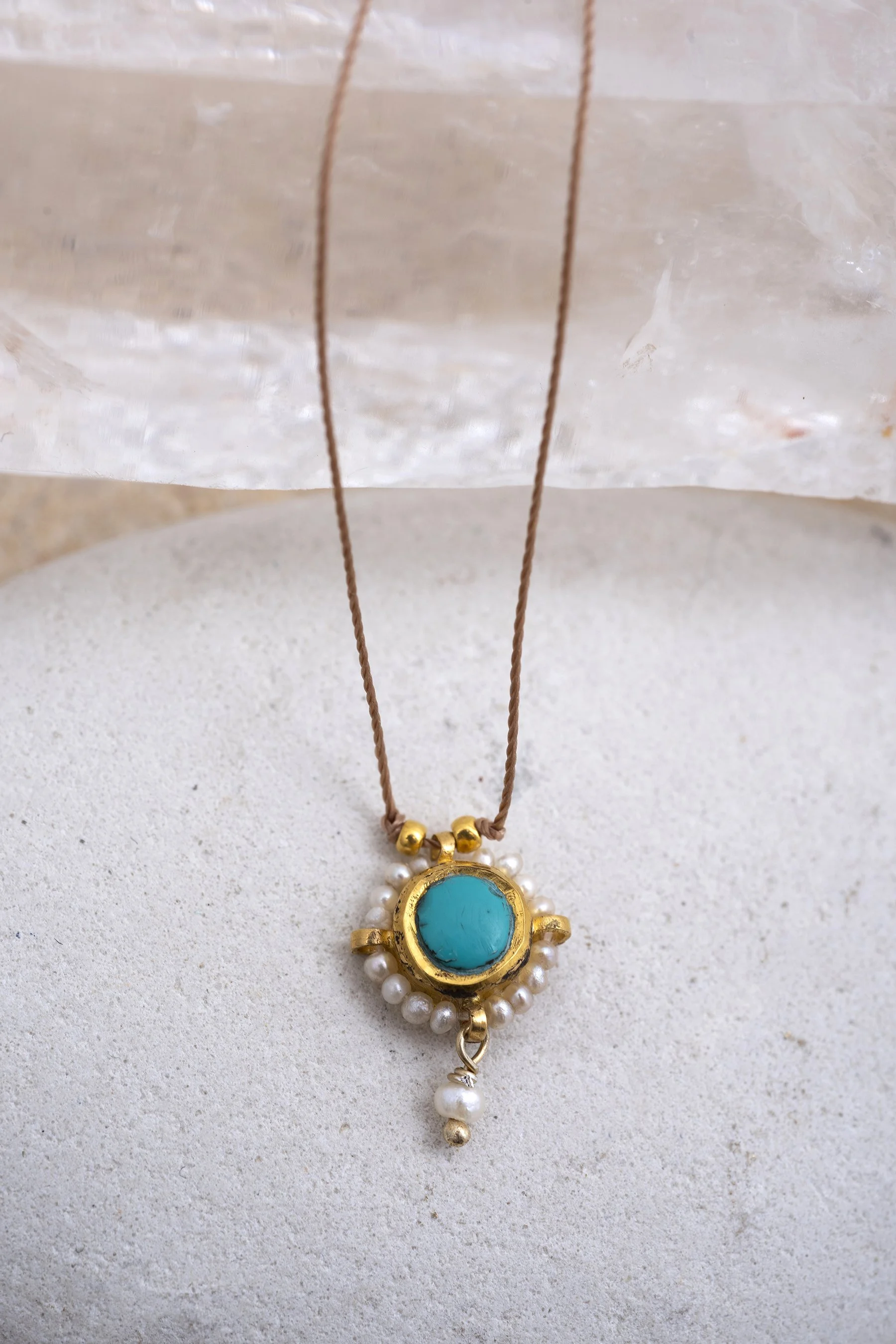 Gold necklace with a turquoise stone pendant surrounded by small pearls, hanging on a beige textured surface.