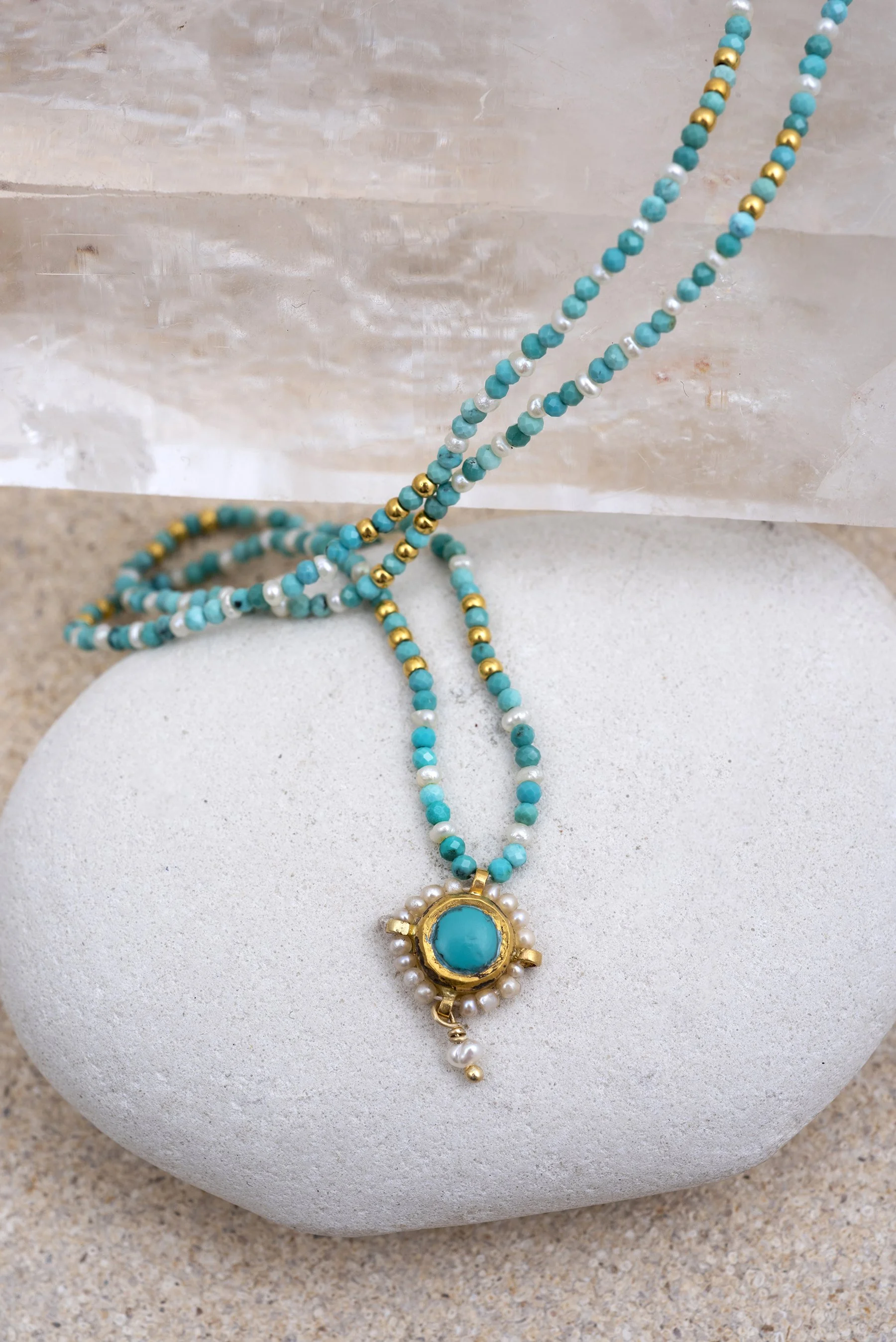 Turquoise, white, and gold beaded necklace with a large turquoise pendant and small pearl accents displayed on a white stone surface with a clear block in the background.