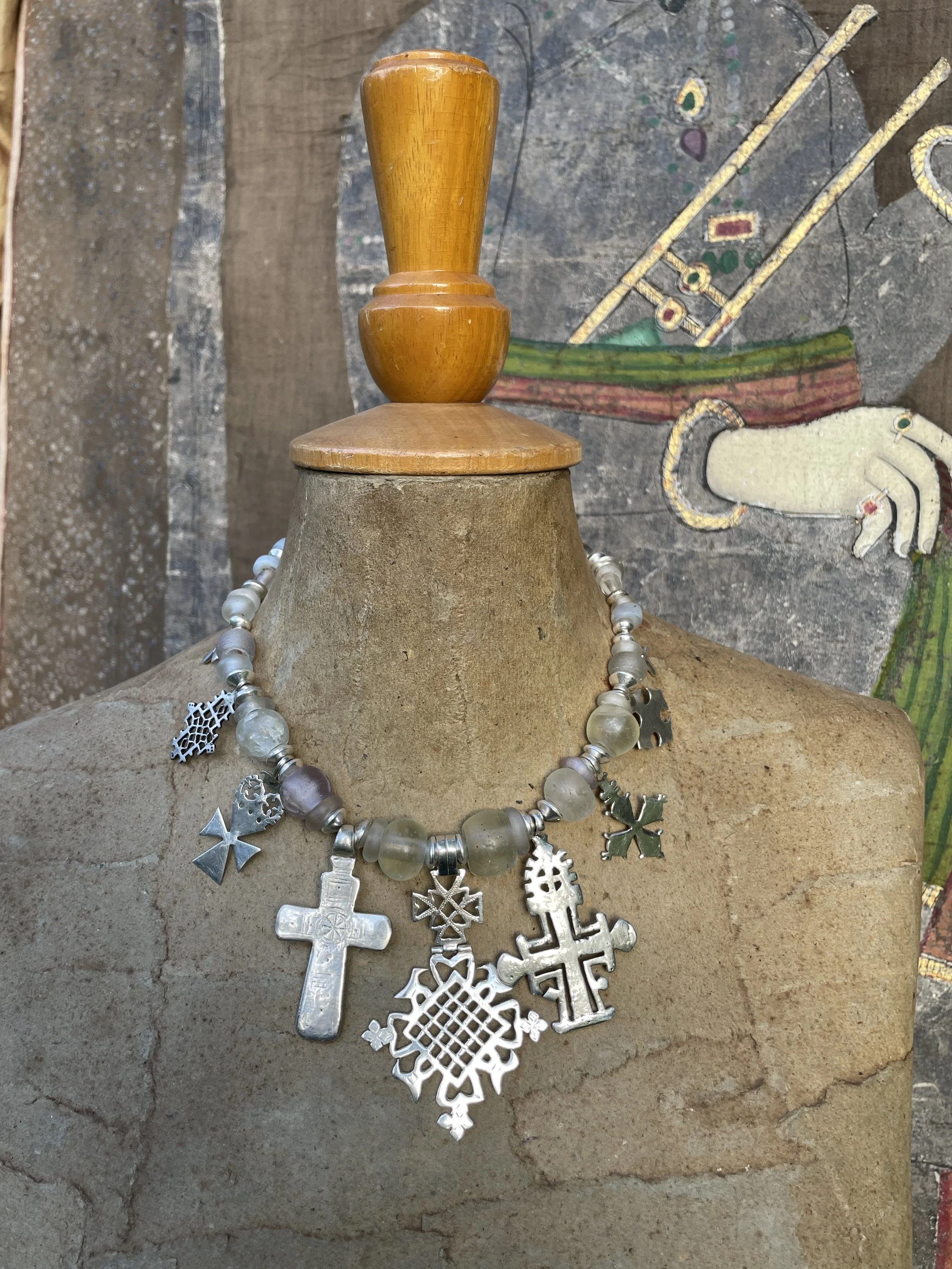 A silver charm necklace displayed on a bust, with multiple cross and other religious charms, resting against a wooden stand with a painted or textured background.