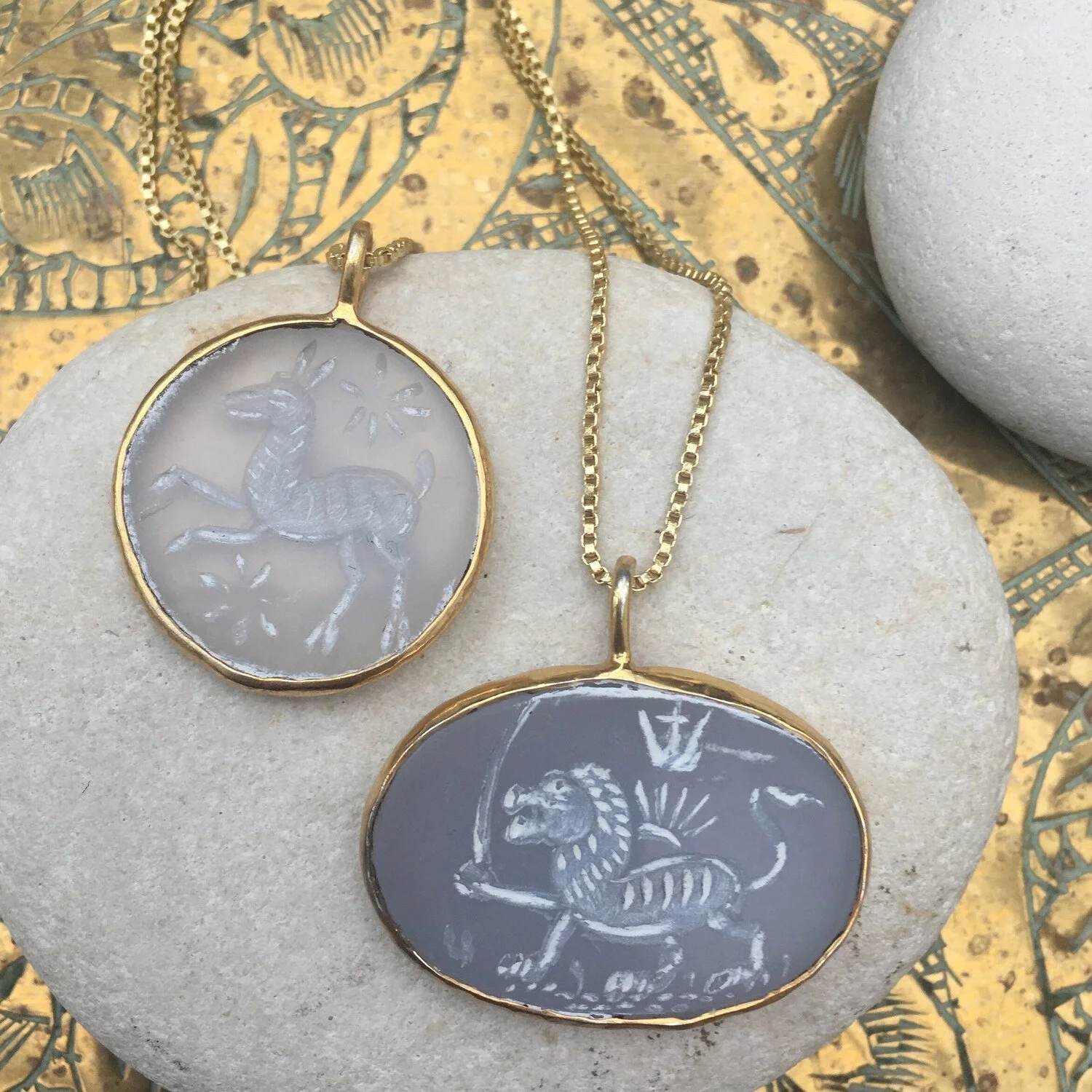 Two gold-framed pendants with engraved animal images, one with a unicorn and the other with a lion, resting on a stone surface.