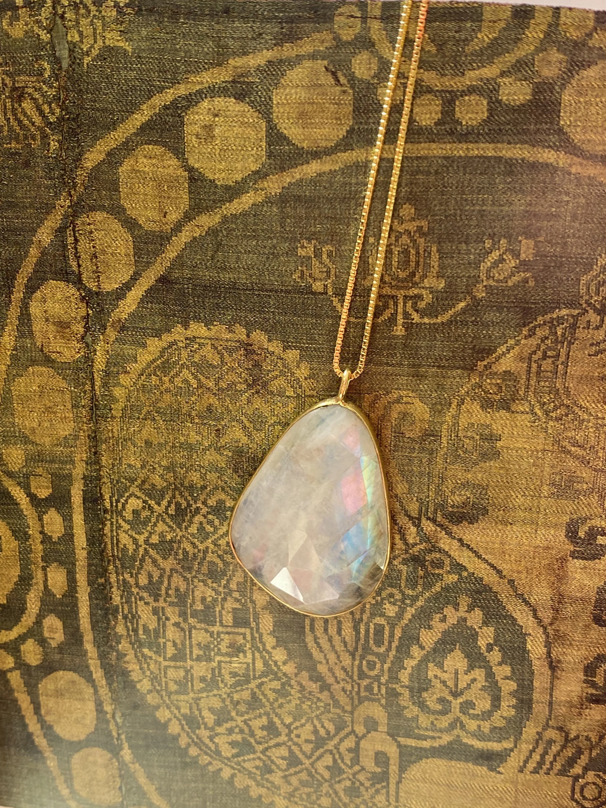 A teardrop-shaped iridescent pendant necklace hanging from a gold chain, resting on a vintage tapestry fabric with floral and geometric patterns.